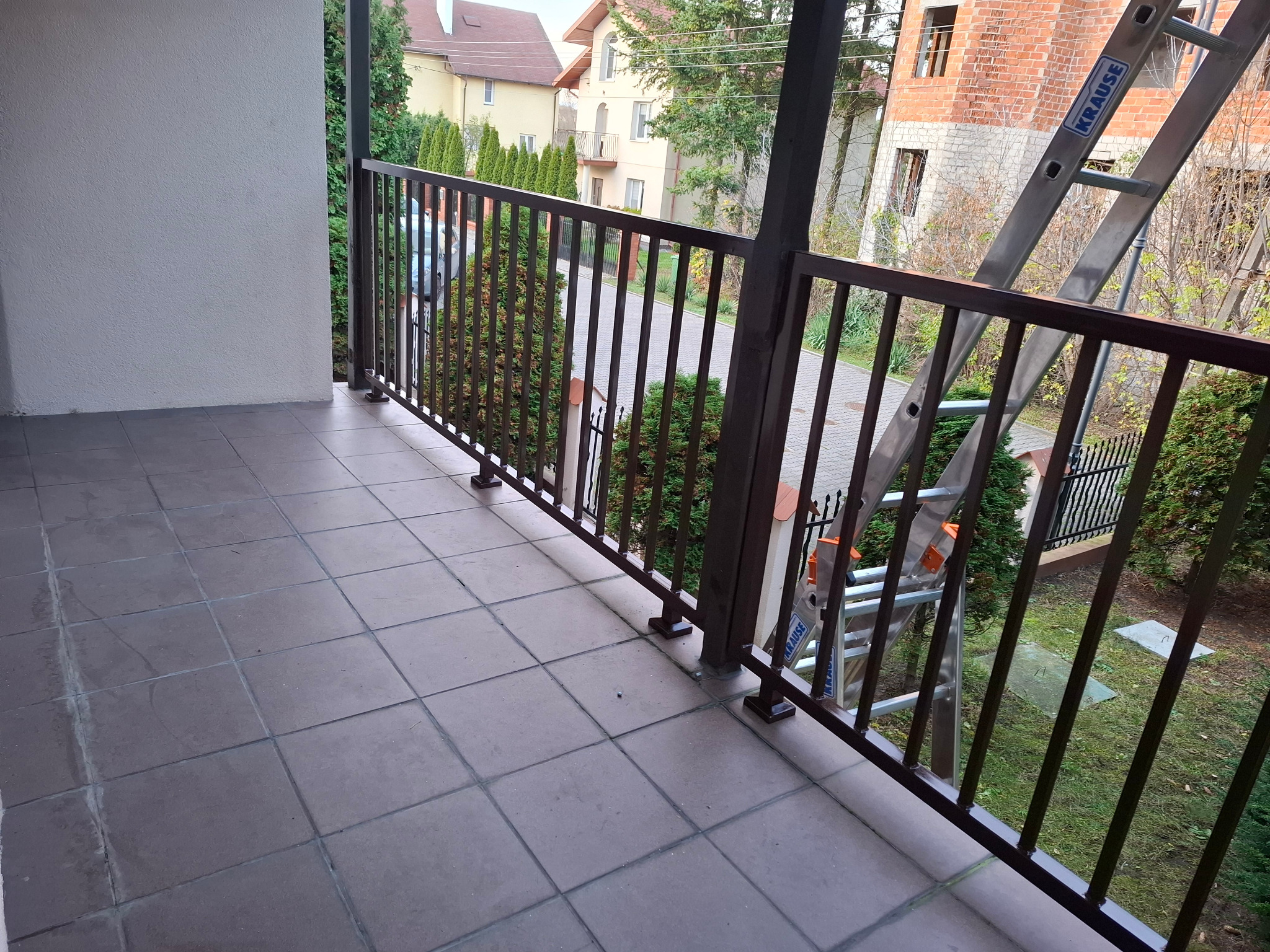 Balkon z metalową balustradą w kolorze ciemnego brązu, widok z góry na kafelki podłogowe i otoczenie z zielenią. W tle budynek w trakcie budowy i drabina.