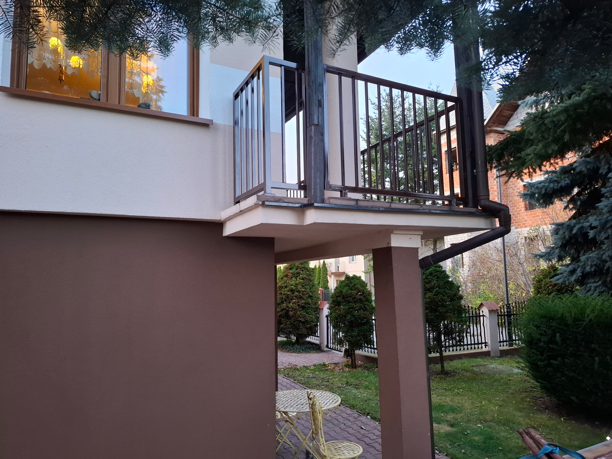 Balkon z brązową, metalową balustradą, widoczny z dołu. Na balkonie widoczne pionowe elementy balustrady. W tle zieleń ogrodu i fragment budynku.