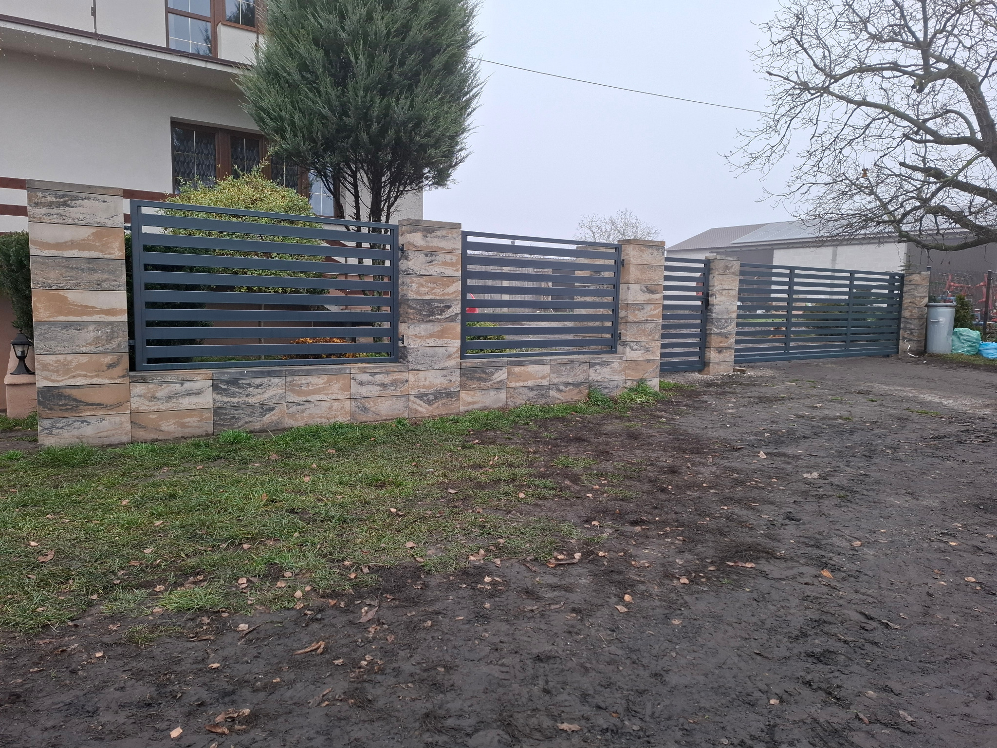 Nowoczesna brama wjazdowa z poziomych, szarych profili, osadzona na słupkach z dekoracyjnej, warstwowej kostki. Tło: budynek i drzewo bez liści.