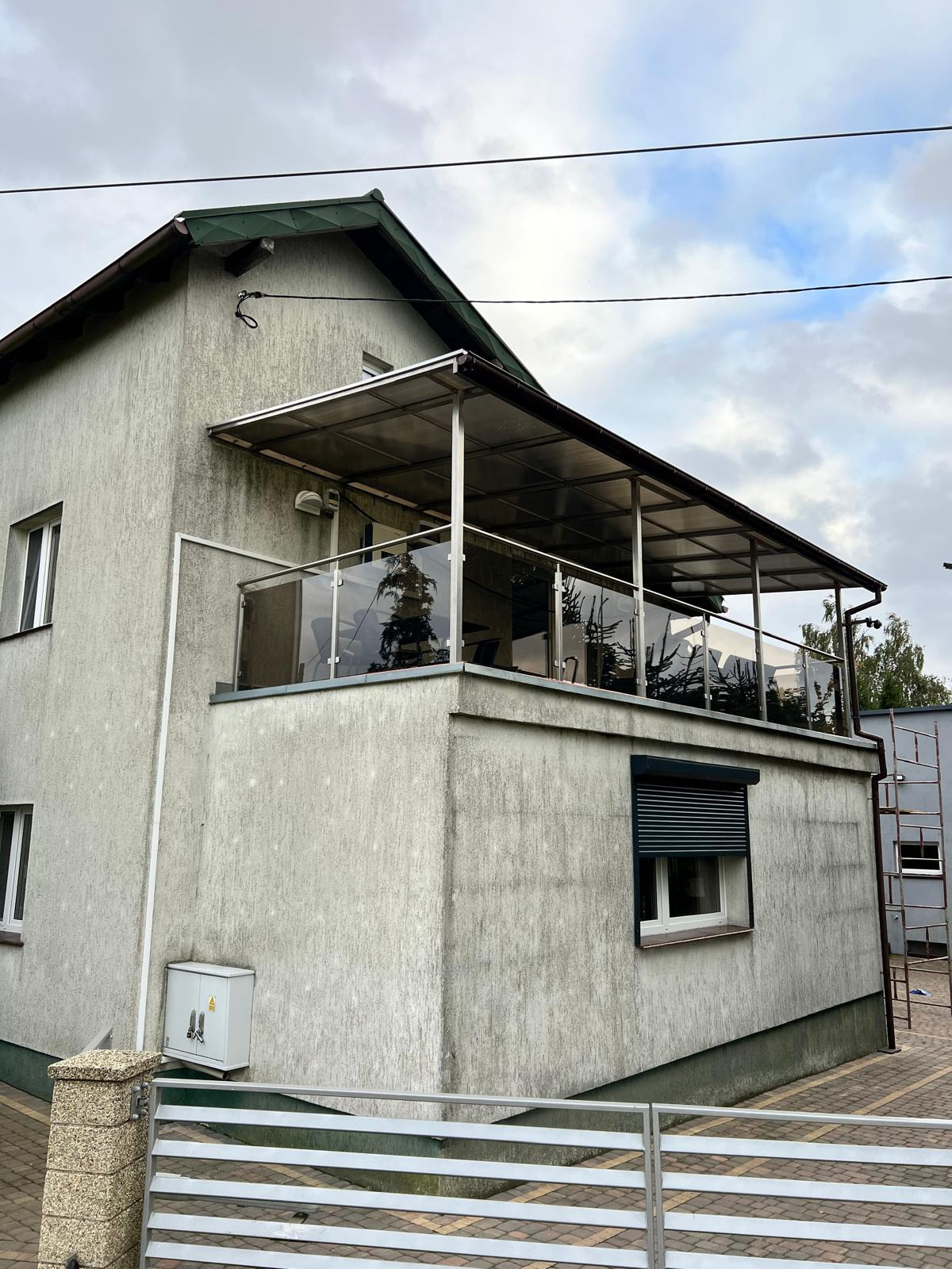 Zadaszony balkon z przeszkloną balustradą na piętrze domu. Widoczne elementy konstrukcyjne i zadaszenie z poliwęglanu. Ujęcie z dołu, pochmurne niebo w tle.