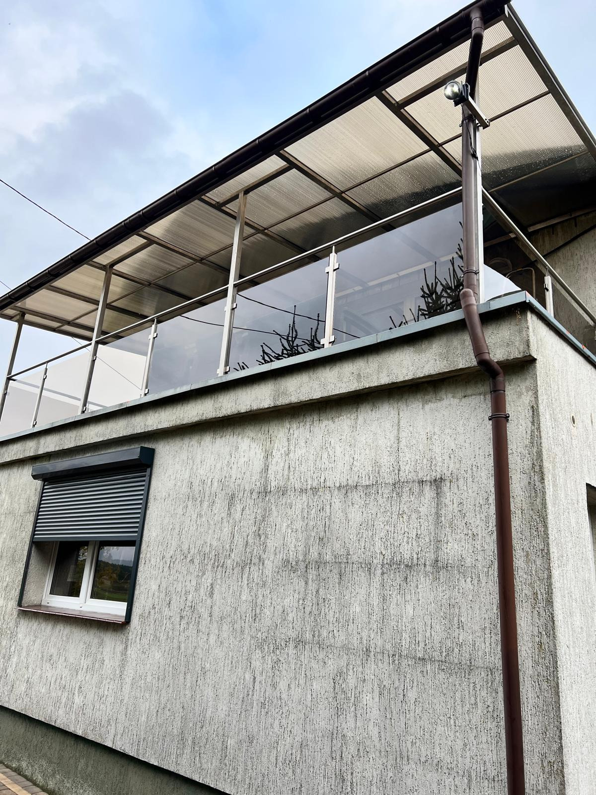 Szklana balustrada balkonowa z zadaszeniem, widok z dołu, z rynną i oknem z roletą. Nowoczesny design z elementami metalowymi.