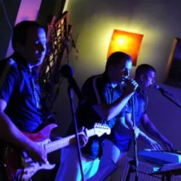 Trzech mężczyzn w koszulach polo grających muzykę na scenie: gitarzysta z czerwoną gitarą, wokalista z mikrofonem i klawiszowiec z keyboardem, oświetleni niebieskim światłem.