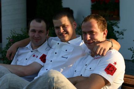 Trzech uśmiechniętych mężczyzn w białych koszulach z czerwonymi naszywkami siedzi obok siebie na ławce, przytuleni ramionami, w tle widoczne krzewy z czerwonymi kwiatami.