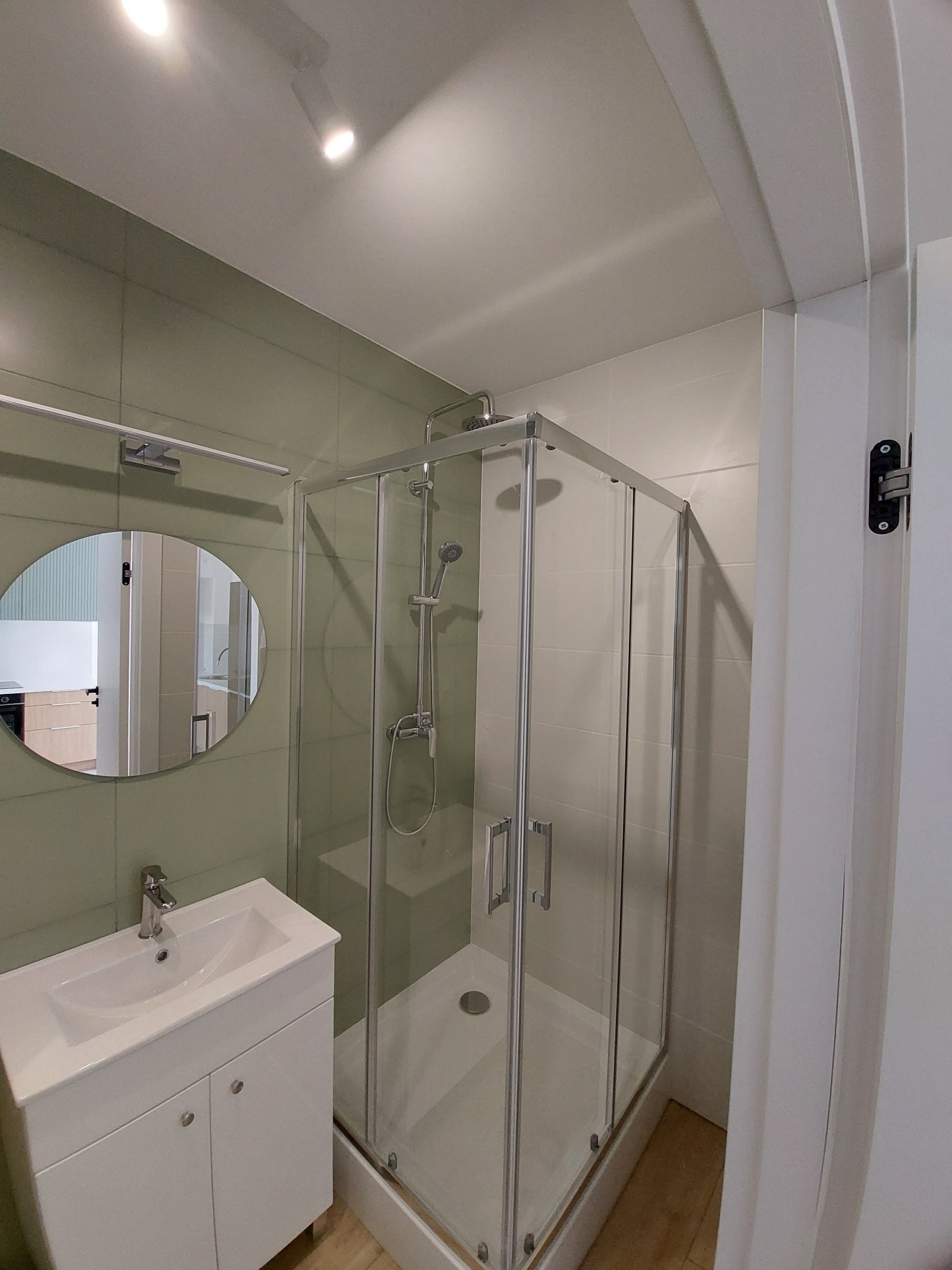 Nowoczesna łazienka z kabiną prysznicową i okrągłym lustrem. Jasne płytki, biała umywalka z szafką. Minimalistyczny design, widok na wnętrze przez lustro.