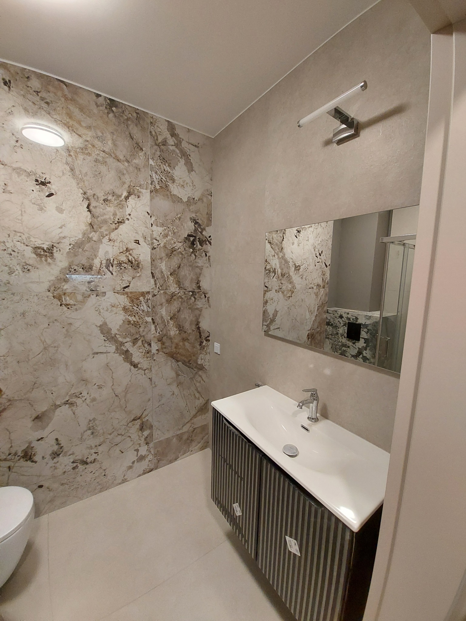 Wykończona łazienka z marmurowymi płytkami na ścianie prysznicowej, umywalką z szafką w paski i lustrem. Nowoczesne oświetlenie i jasne płytki podłogowe.