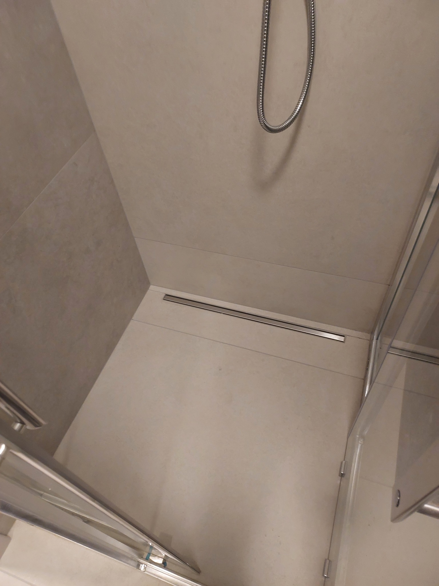 Nowoczesny prysznic walk-in z odpływem liniowym i szklaną kabiną. Beżowe płytki tworzą minimalistyczny design. Widoczny wąż prysznicowy.