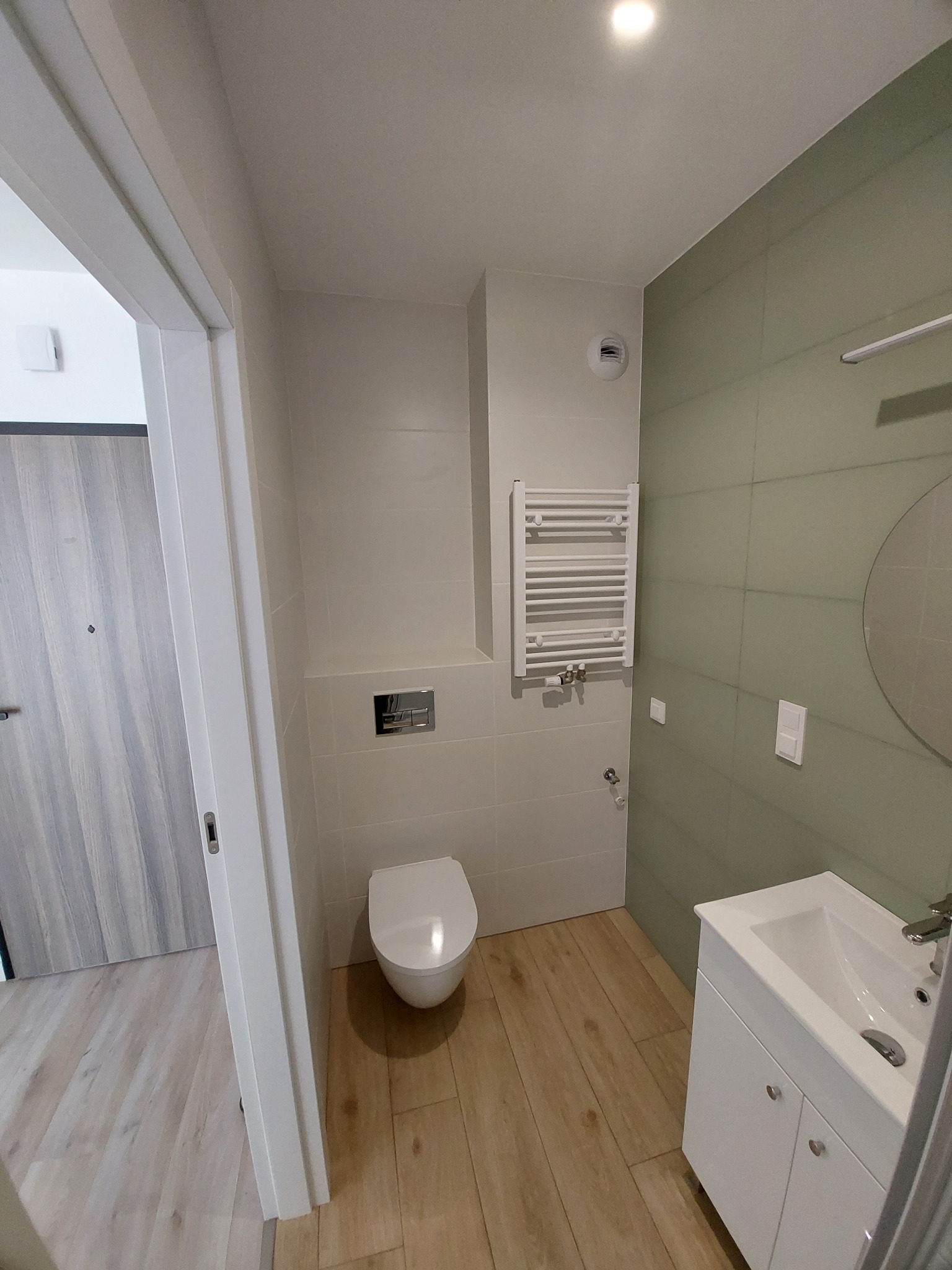 Jasna łazienka z drewnopodobną podłogą, umywalką z szafką, toaletą i grzejnikiem drabinkowym. Ściany wyłożone płytkami w odcieniach bieli i zieleni. Minimalistyczny styl.