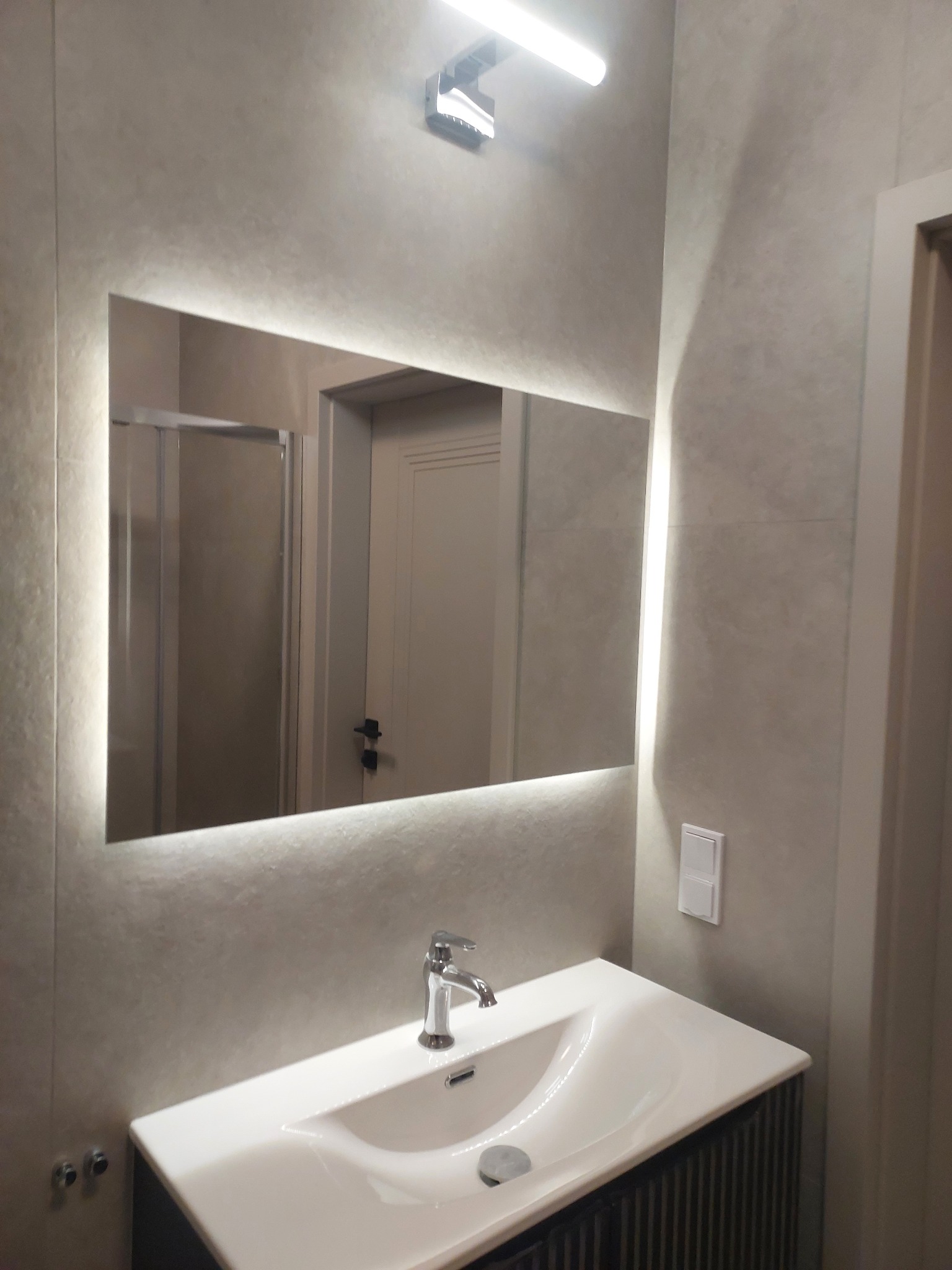 Nowoczesna łazienka: umywalka z szafką, podświetlane lustro i minimalistyczna armatura na tle płytek w odcieniach szarości. Widoczny fragment kabiny prysznicowej w odbiciu.