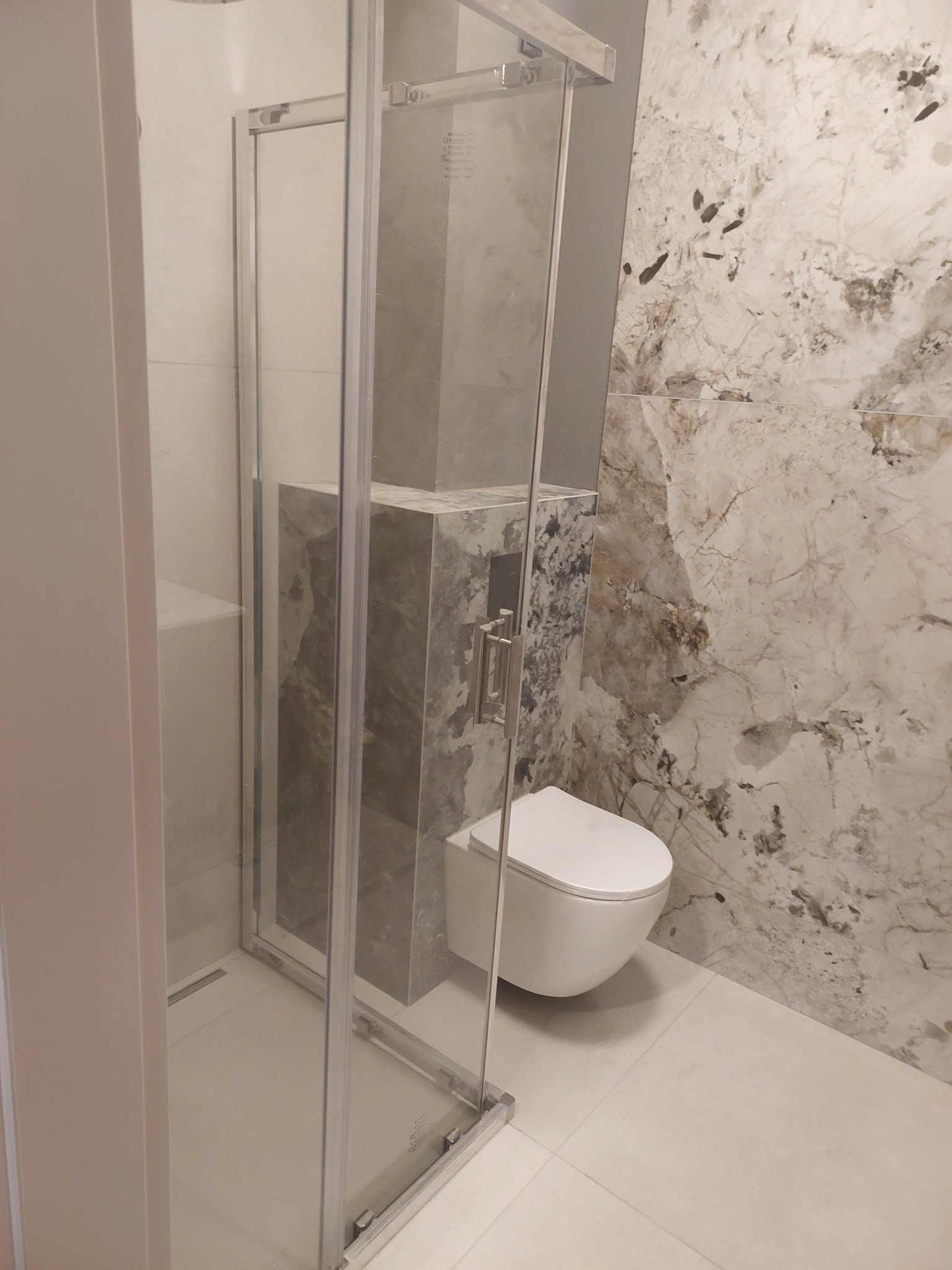 Nowoczesna łazienka z kabiną prysznicową typu walk-in, podwieszaną toaletą i marmurowymi płytkami na ścianie. Minimalistyczny design z akcentem na naturalne materiały.