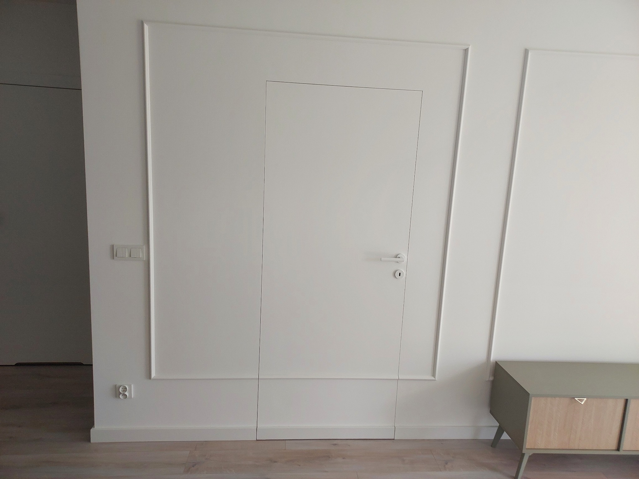 Białe, ukryte drzwi w ścianie z dekoracyjnymi listwami, minimalistyczny design, widoczny fragment podłogi i szafki. Nowoczesne wykończenie wnętrza.