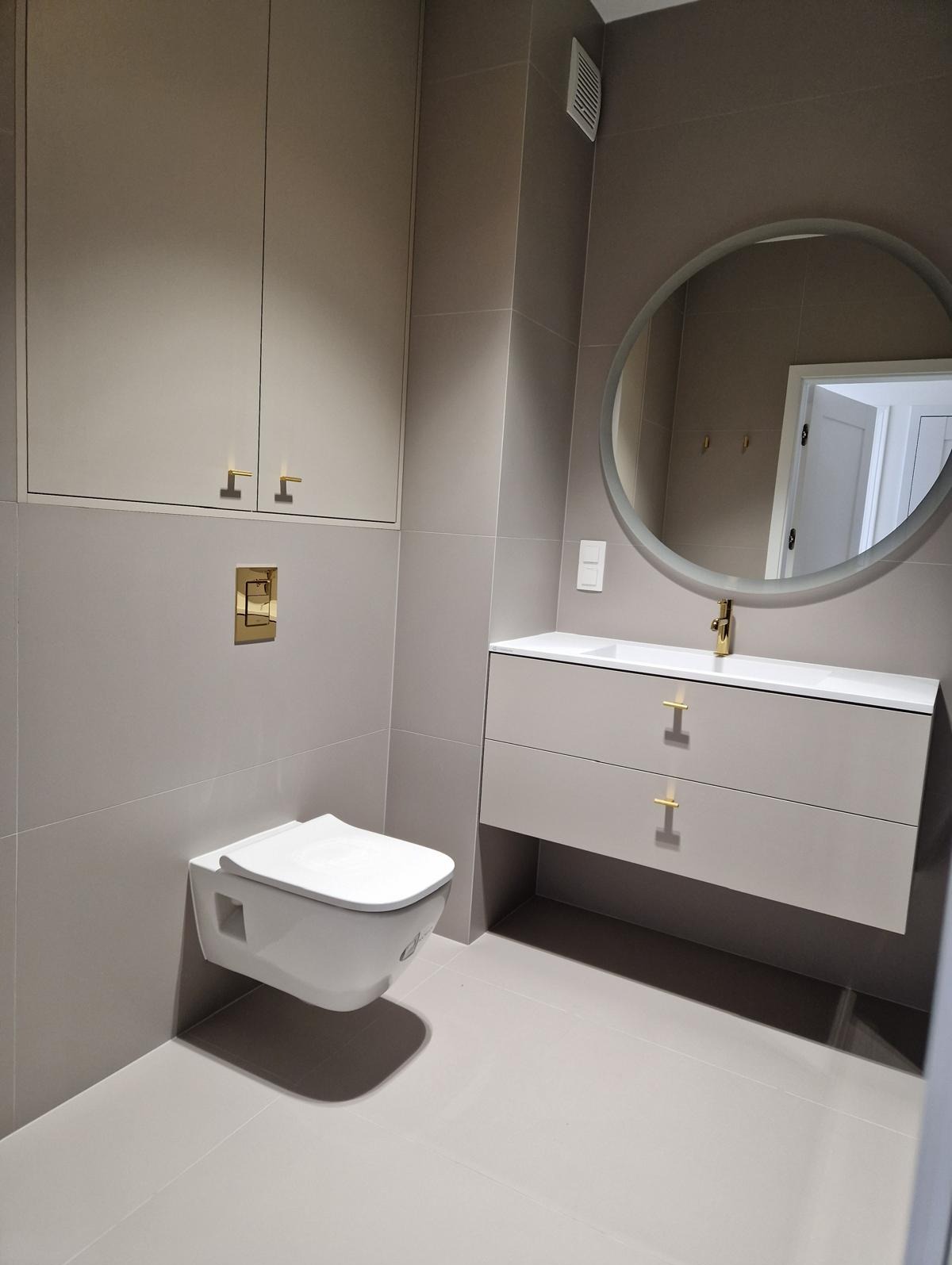 Nowoczesna łazienka w odcieniach szarości z wiszącą toaletą, szafką z umywalką i okrągłym lustrem. Złote akcenty armatury i uchwytów dodają elegancji.
