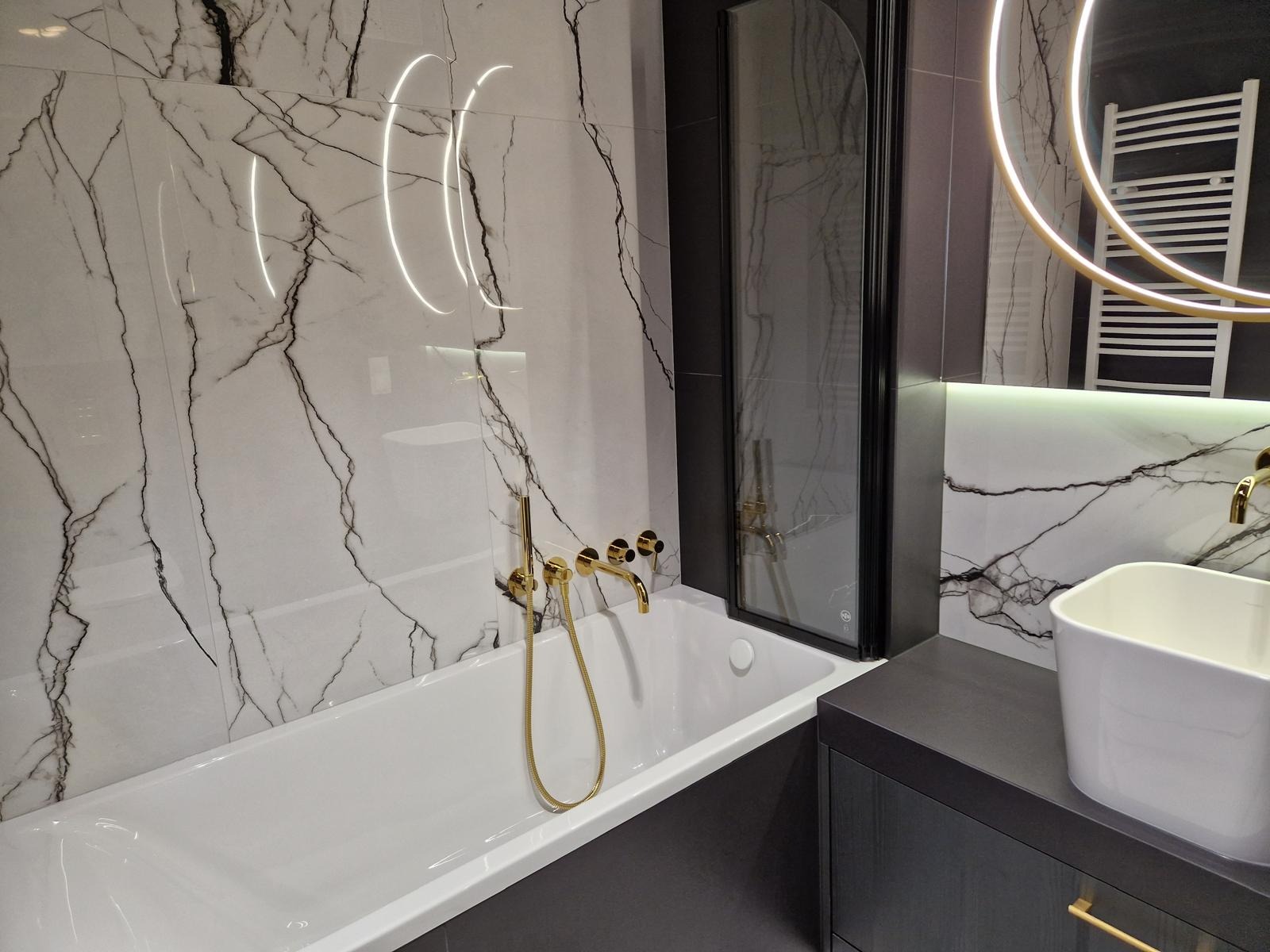 Elegancka łazienka po remoncie: białe marmurowe płytki z czarnymi żyłami, złota armatura, czarna kabina prysznicowa i okrągłe lustro z podświetleniem LED.