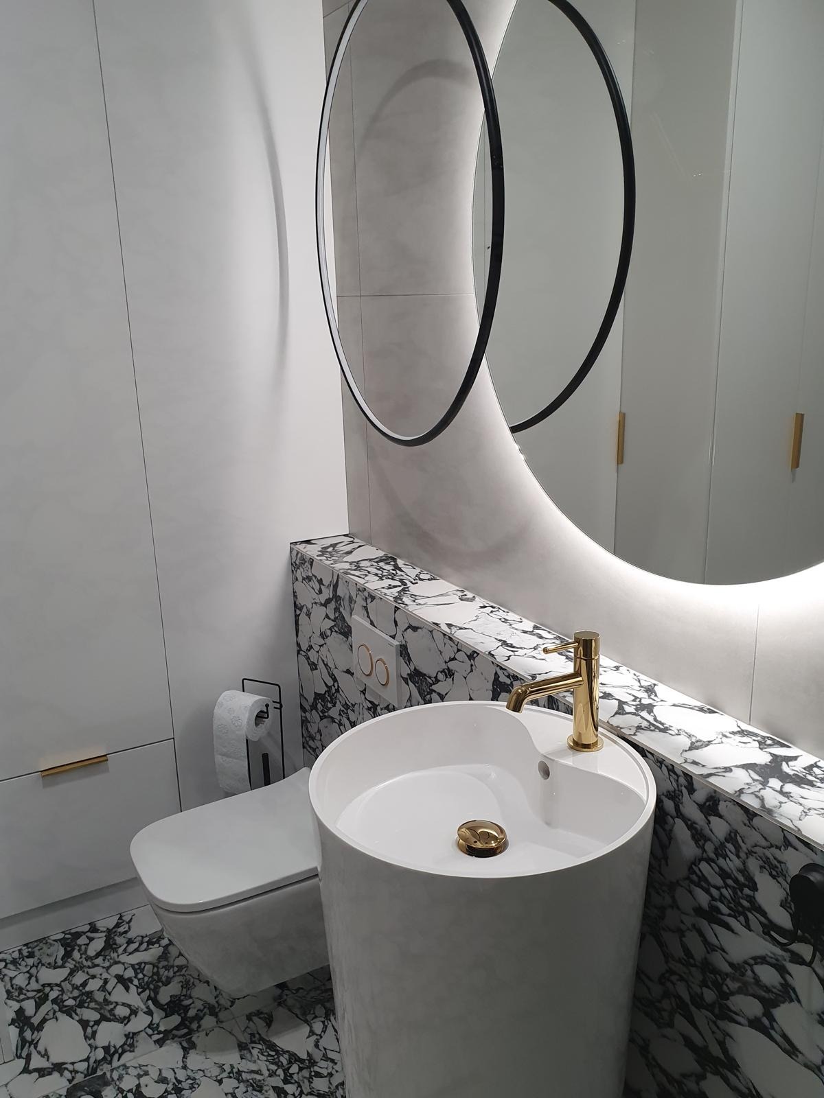 Nowoczesna łazienka z okrągłym lustrem z podświetleniem LED, białą umywalką na postumencie i marmurowym wykończeniem ściany. Złota bateria i minimalistyczna toaleta.