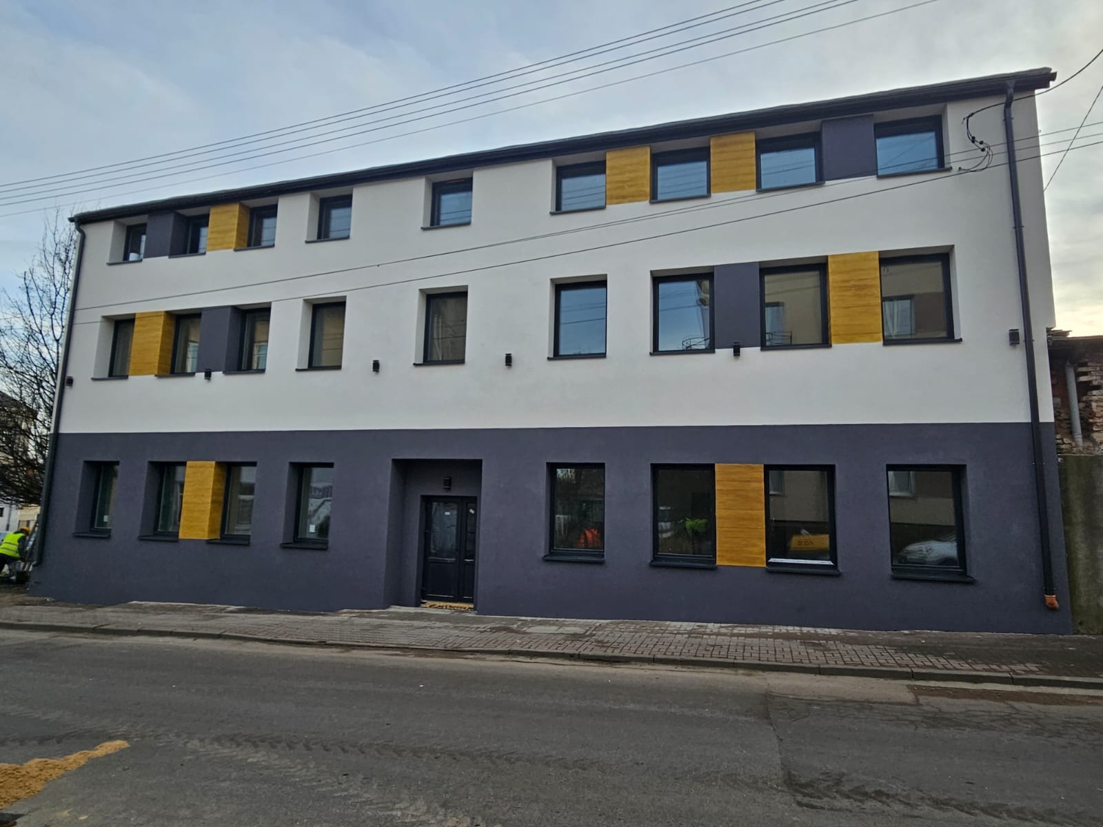 Odnowiona kamienica po termomodernizacji z nowoczesną elewacją w odcieniach szarości, bieli i akcentami drewna. Widok z poziomu ulicy, pochmurne niebo.