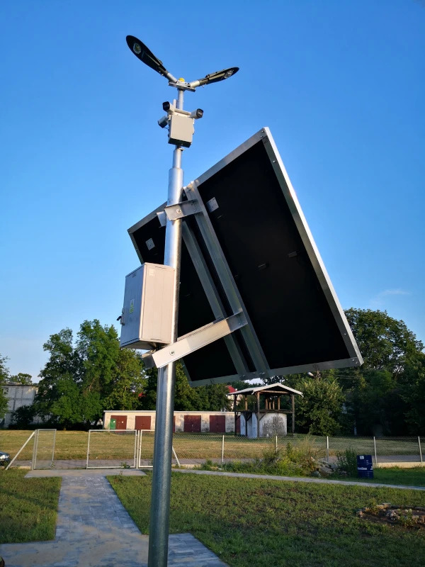 Lampa solarna z panelem fotowoltaicznym na tle błękitnego nieba i zieleni. Widoczny metalowy słup, oświetlenie LED i skrzynka sterownicza. Ujęcie z dołu.