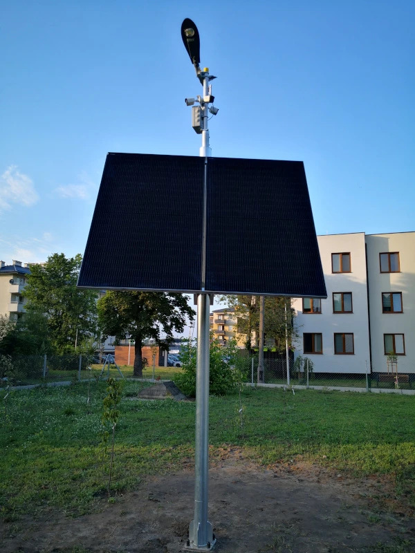 Lampa solarna z panelem fotowoltaicznym i kamerami monitoringu na tle budynku mieszkalnego i zieleni miejskiej, widok z dołu.