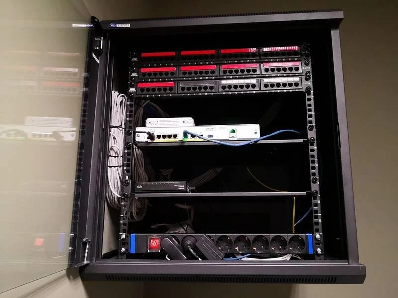 Szafa rack z zainstalowanymi urządzeniami sieciowymi: patch panel, routery, listwa zasilająca. Widoczne okablowanie i elementy montażowe. Otwarta szyba szafy.