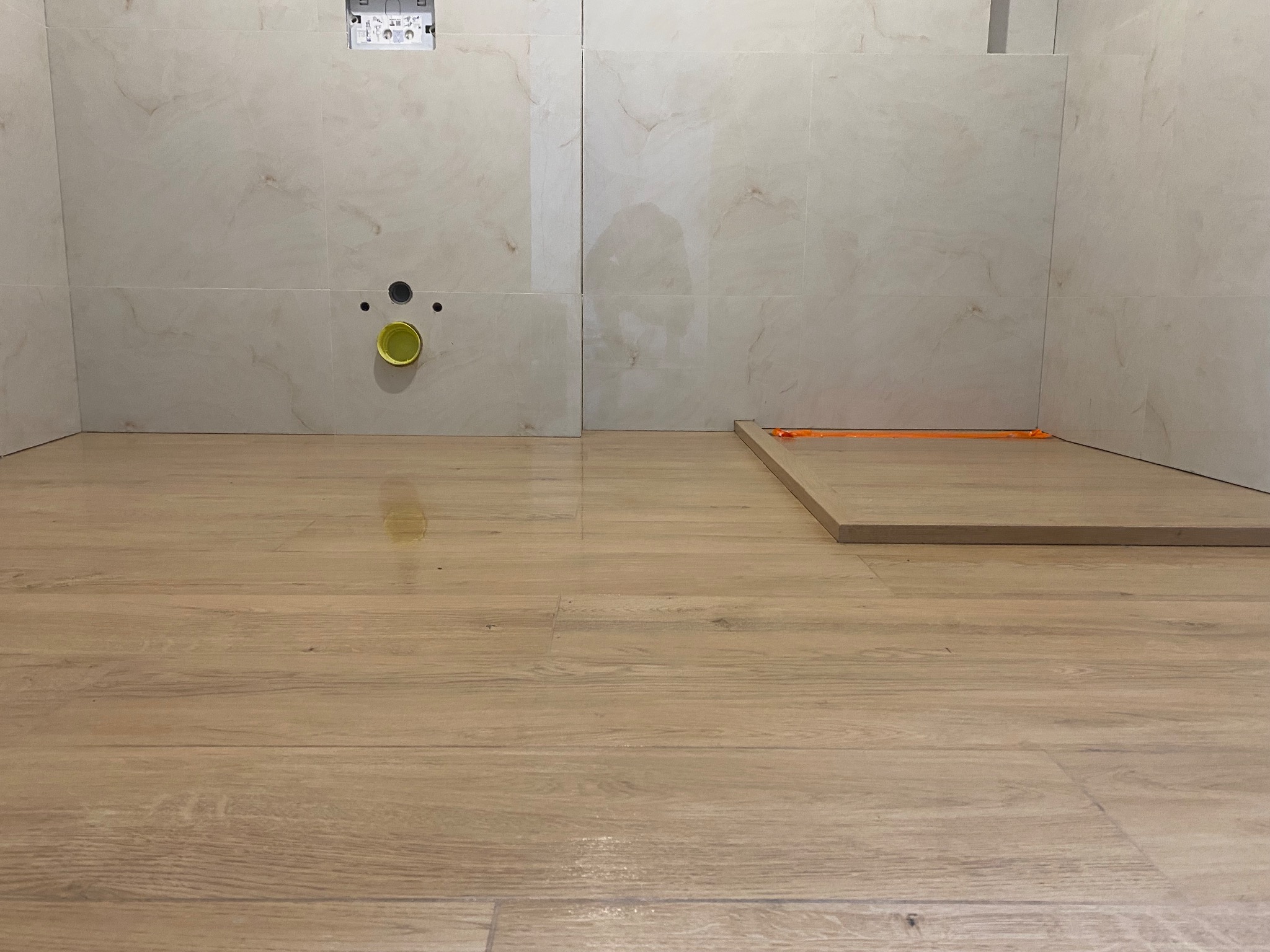 Jasne płytki imitujące marmur na ścianie i drewnopodobna podłoga w łazience. Widoczne elementy instalacji i fragment płyty podłogowej z pomarańczowymi podkładkami.