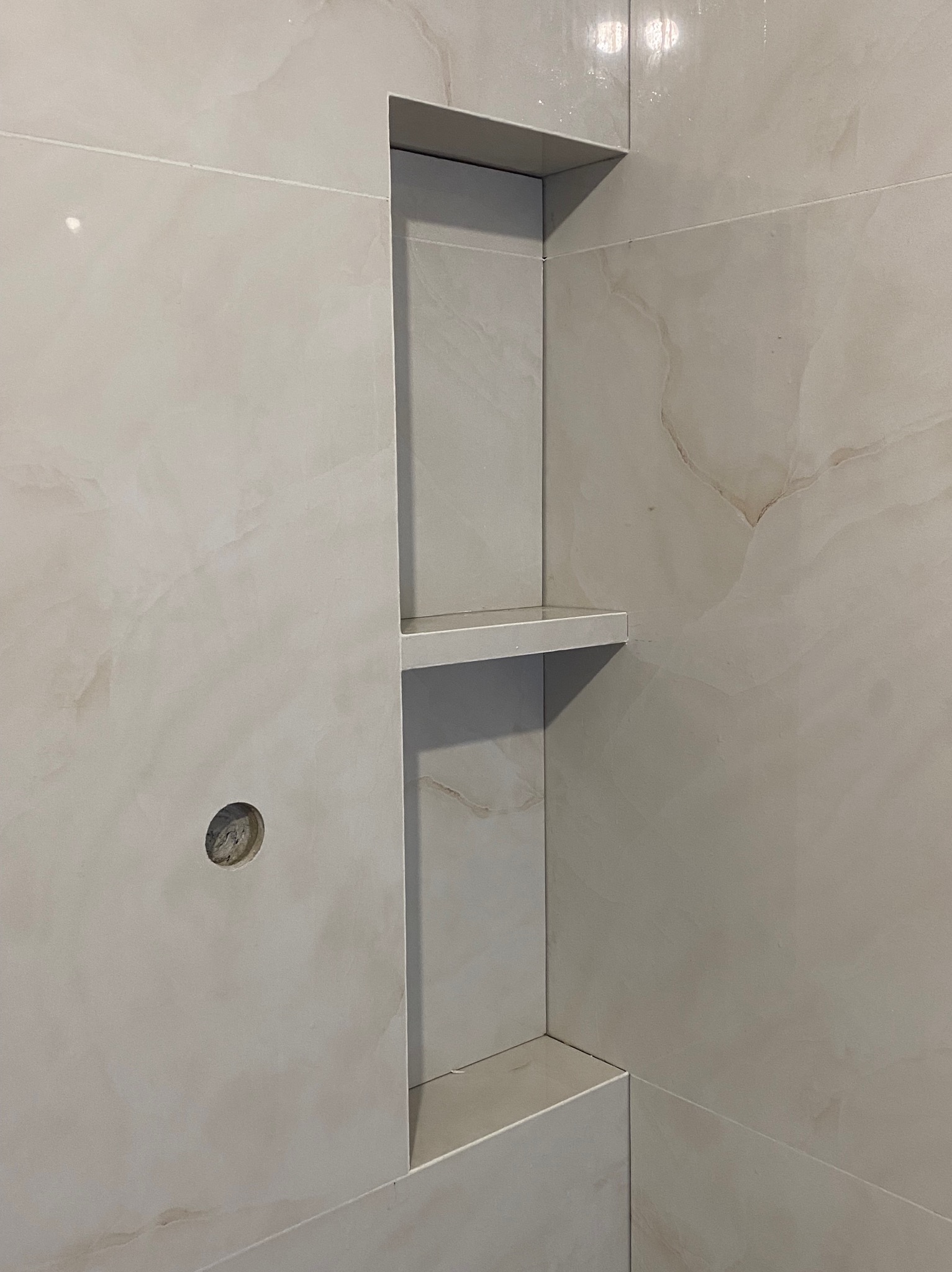 Beżowa, marmurkowa ściana łazienki z wnęką i półkami wykończonymi płytkami. Widoczny otwór na instalację. Minimalistyczny design, ciepłe oświetlenie.