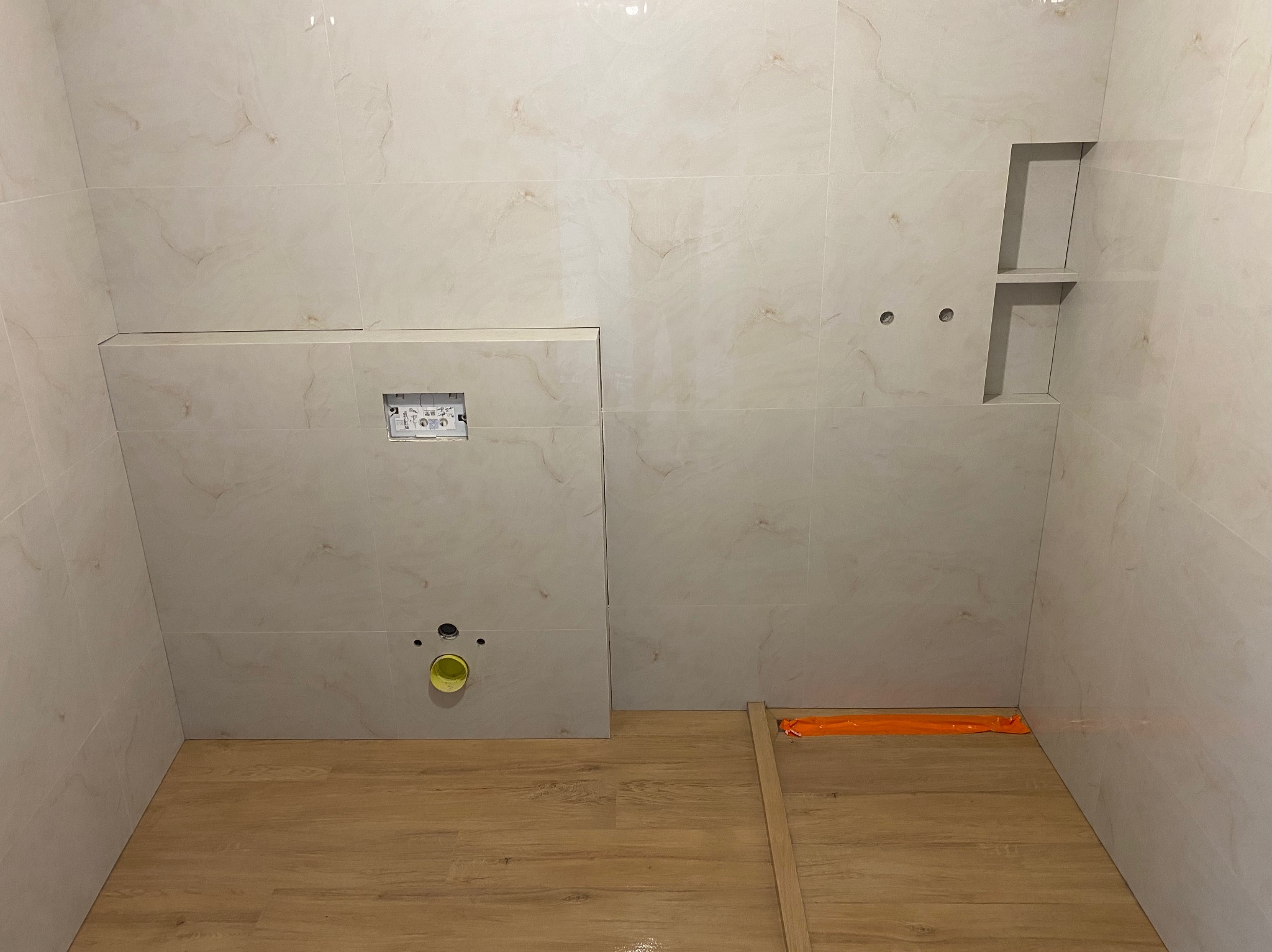 Wykończona łazienka z beżowymi płytkami imitującymi marmur, drewnopodobną podłogą i wnęką z półkami. Widoczne otwory na armaturę i stelaż podtynkowy WC.