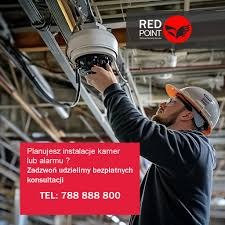 Instalator w kasku montuje kamerę przemysłową pod sufitem hali. Widoczne kable i rury instalacyjne. Logo Red Point w prawym górnym rogu.