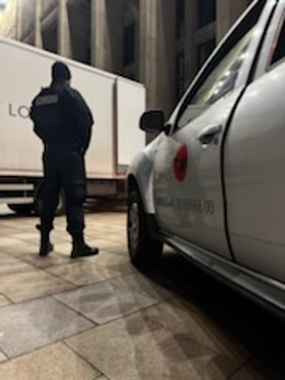 Mężczyzna w uniformie stoi obok białego samochodu z logo firmy ochroniarskiej i białej naczepy na tle kolumn budynku. Nocne oświetlenie.