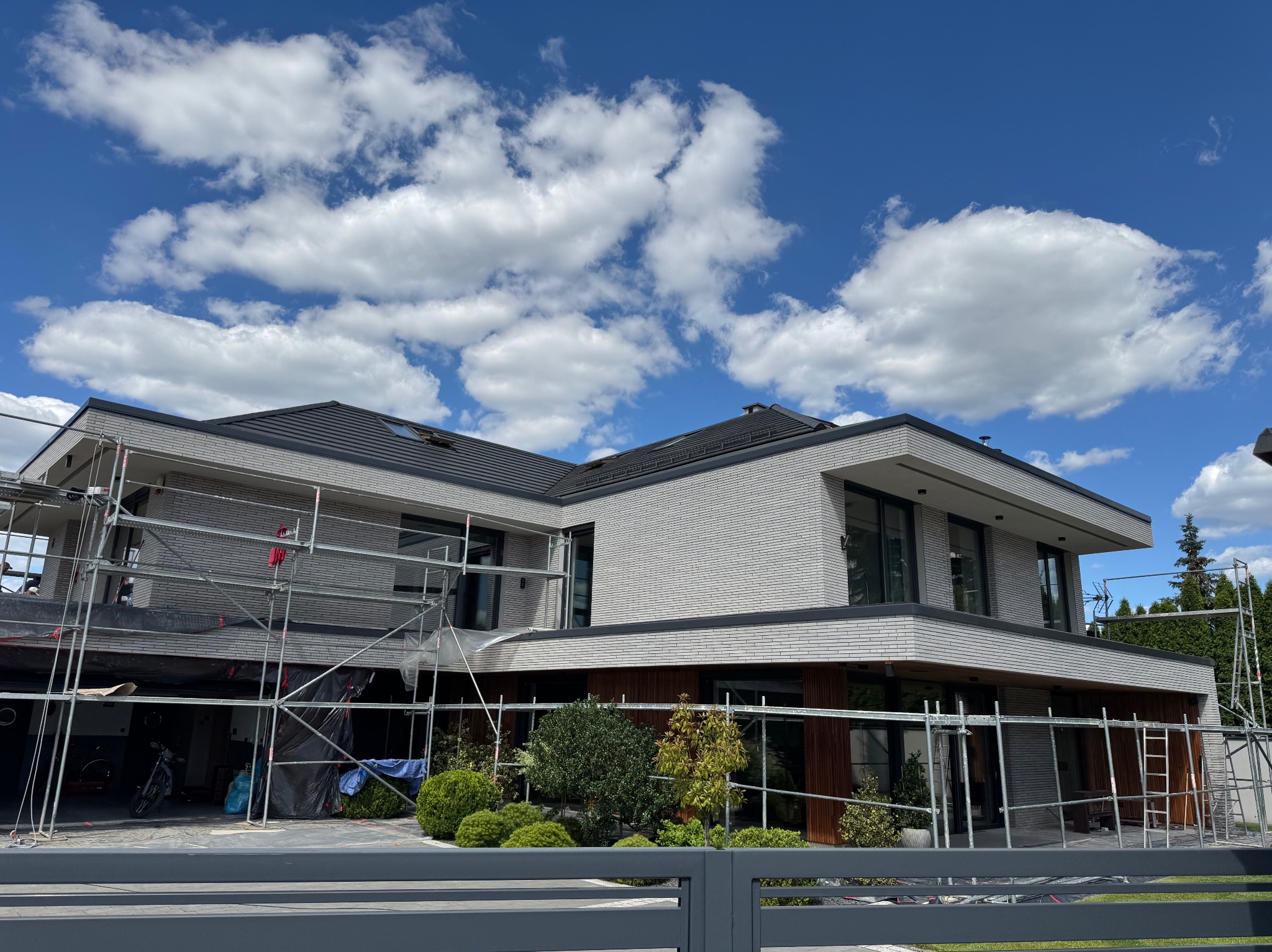 Nowoczesny dom z jasną elewacją w trakcie prac wykończeniowych, widoczne rusztowania. Błękitne niebo z chmurami w tle. Ujęcie zza szarego ogrodzenia.