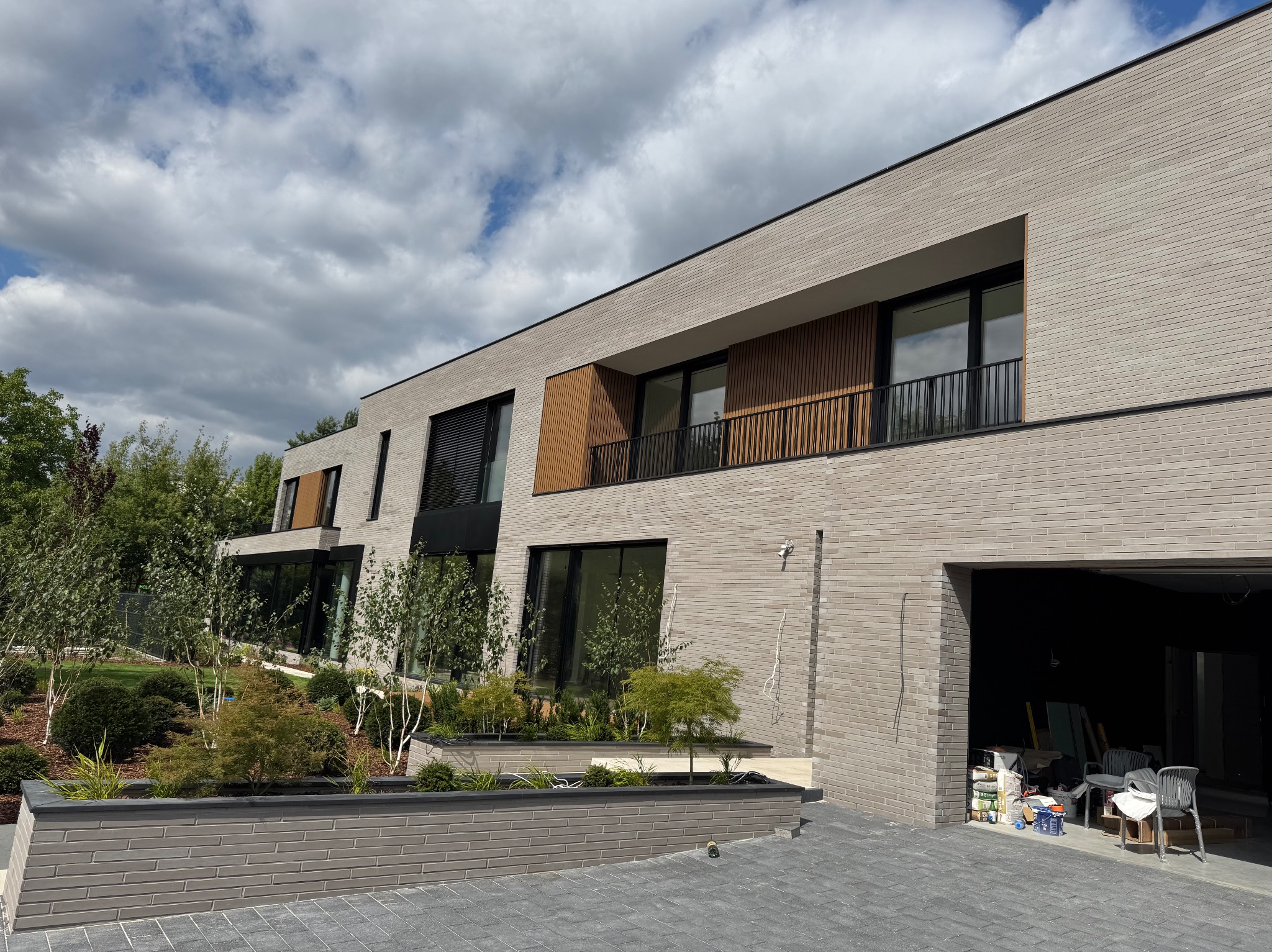 Nowoczesny dom z elewacją z cegły klinkierowej w odcieniach szarości, z drewnianymi elementami dekoracyjnymi i dużymi oknami. Widoczne rośliny w donicach i otwarty garaż.