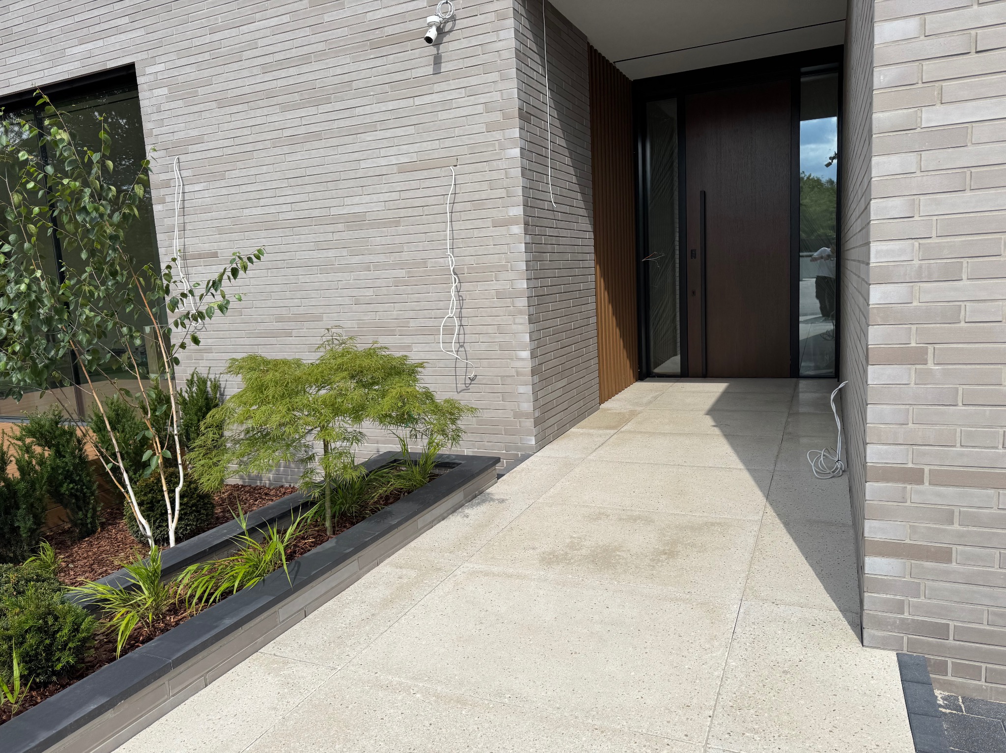 Elegancka elewacja z jasnej cegły klinkierowej, nowoczesne drzwi wejściowe z drewnianymi panelami, minimalistyczny ogródek z roślinami i monitoring.