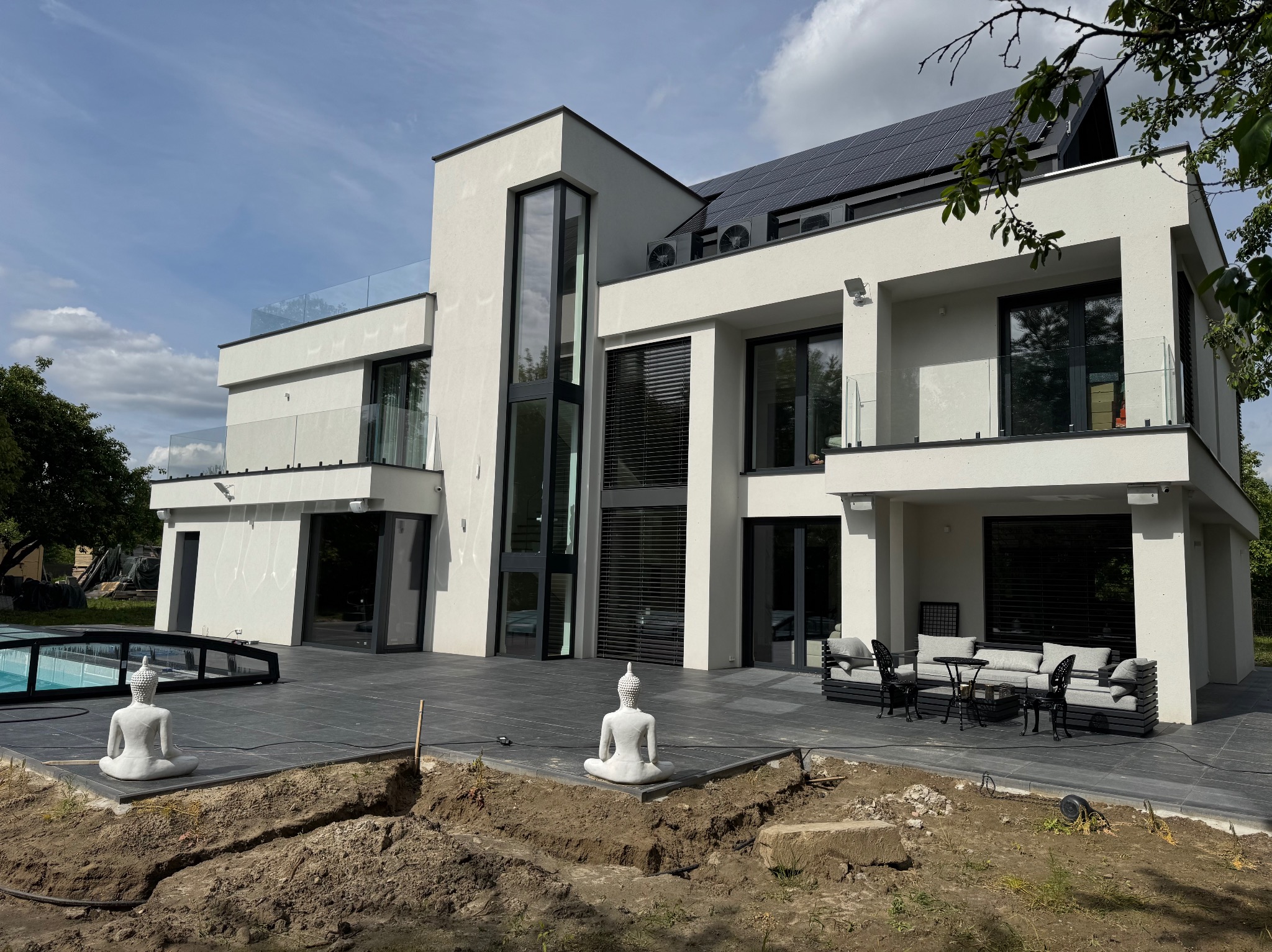 Nowoczesny dom z białą elewacją, panelami słonecznymi i tarasem z posągami Buddy. Widoczne prace ziemne w ogrodzie.