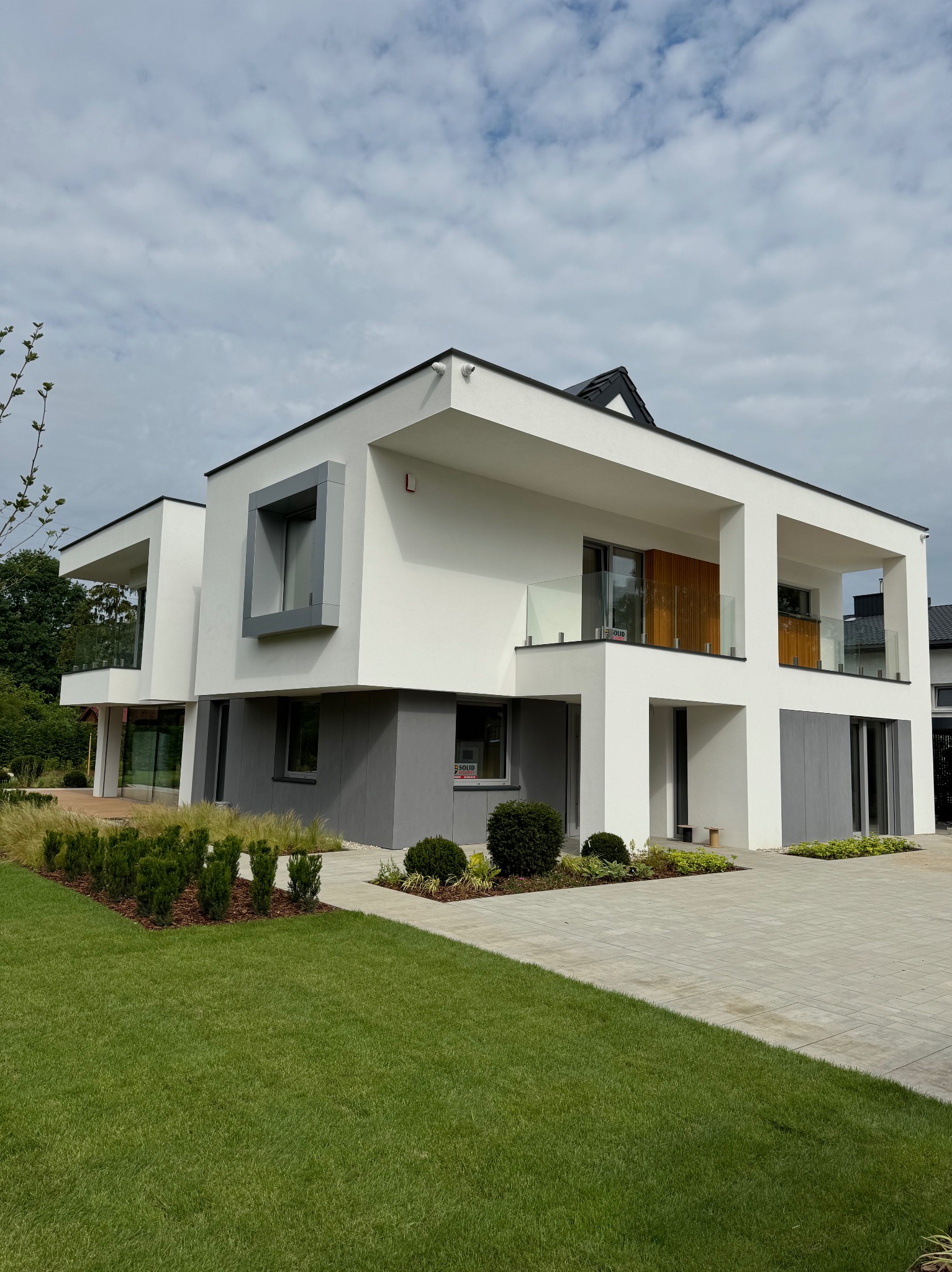 Nowoczesny dom z biało-szarą elewacją, szklanymi balustradami balkonów i zadbanym trawnikiem. Widok z poziomu gruntu, podkreślający minimalistyczny design.