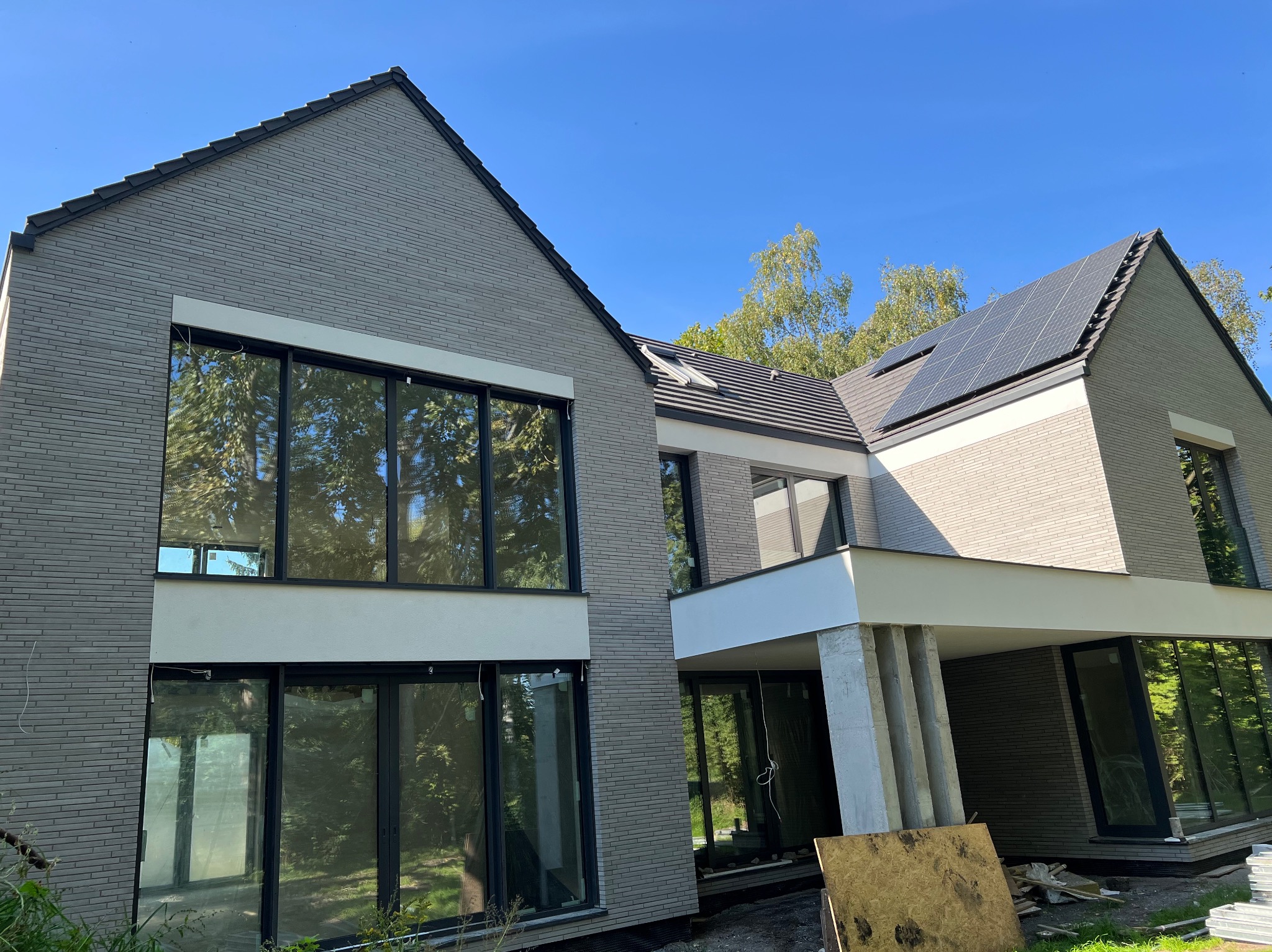 Nowoczesny dom z szarej cegły klinkierowej, duże okna, panele słoneczne na dachu. Widoczne elementy wykończeniowe i fragment terenu wokół budynku.