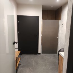 TRU Marcin Dudek - Nowoczesna łazienka z szarą podłogą, czarnymi drzwiami i kabiną prysznicową ze szkła matowego. Widoczna szafka z drewnianym frontem i elementami wyposażenia.