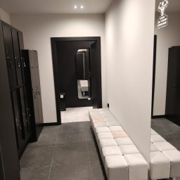 TRU Marcin Dudek - Nowoczesna szatnia w siłowni: czarne szafki, szara podłoga, biała ławka z pufami i lustro z logo 'Gym of Your Dreams'. Minimalistyczny design, oświetlenie punktowe.
