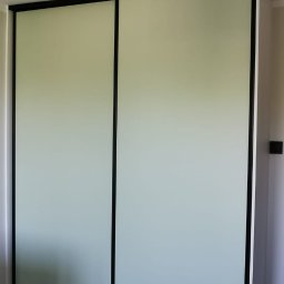 TRU Marcin Dudek - Nowoczesna szafa przesuwna z jasnymi frontami i czarnymi obramowaniami, dopasowana do wnętrza z panelami podłogowymi. Minimalistyczny design.