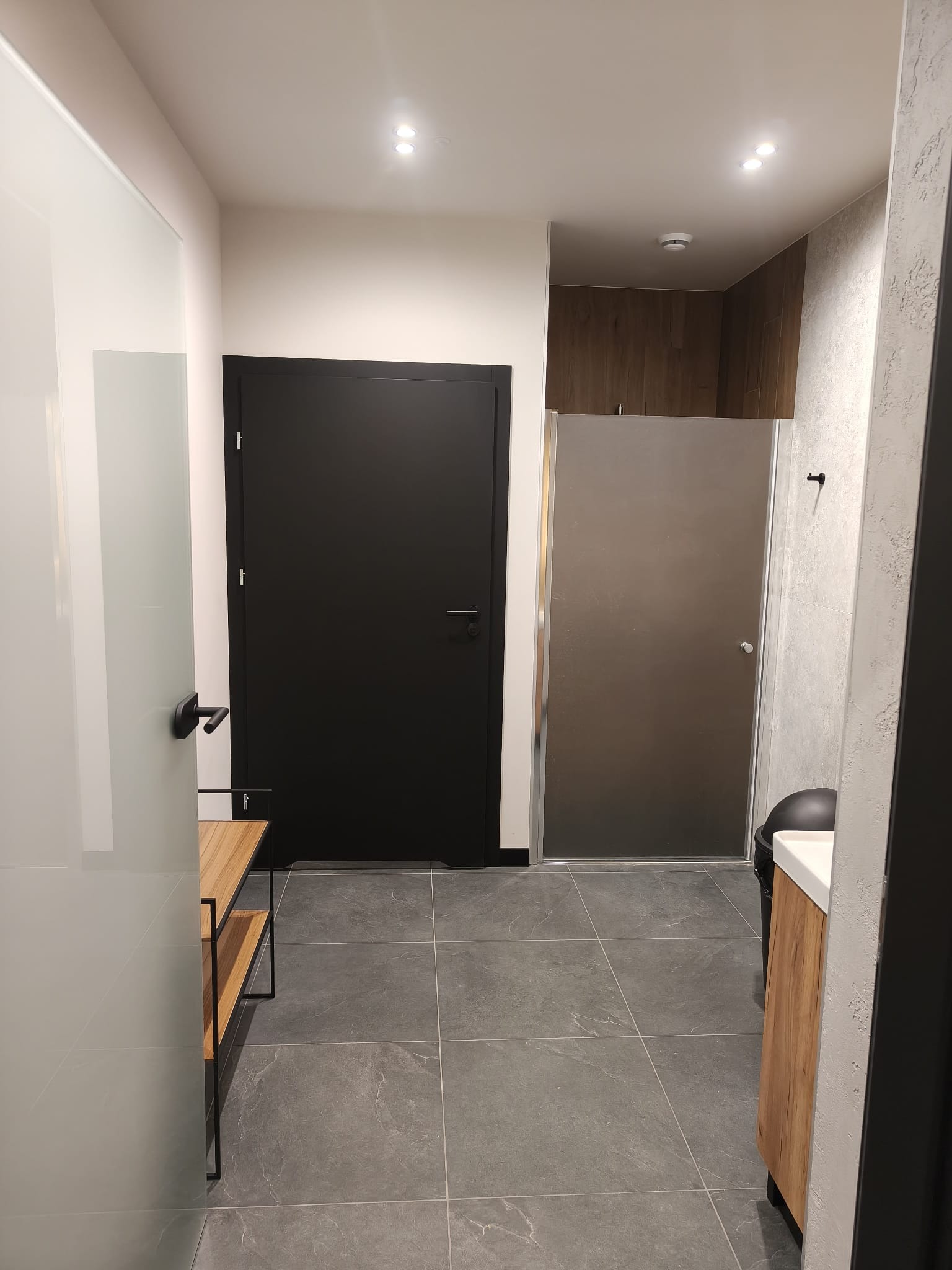 Nowoczesna łazienka z szarą podłogą, czarnymi drzwiami i kabiną prysznicową ze szkła matowego. Widoczna szafka z drewnianym frontem i elementami wyposażenia.