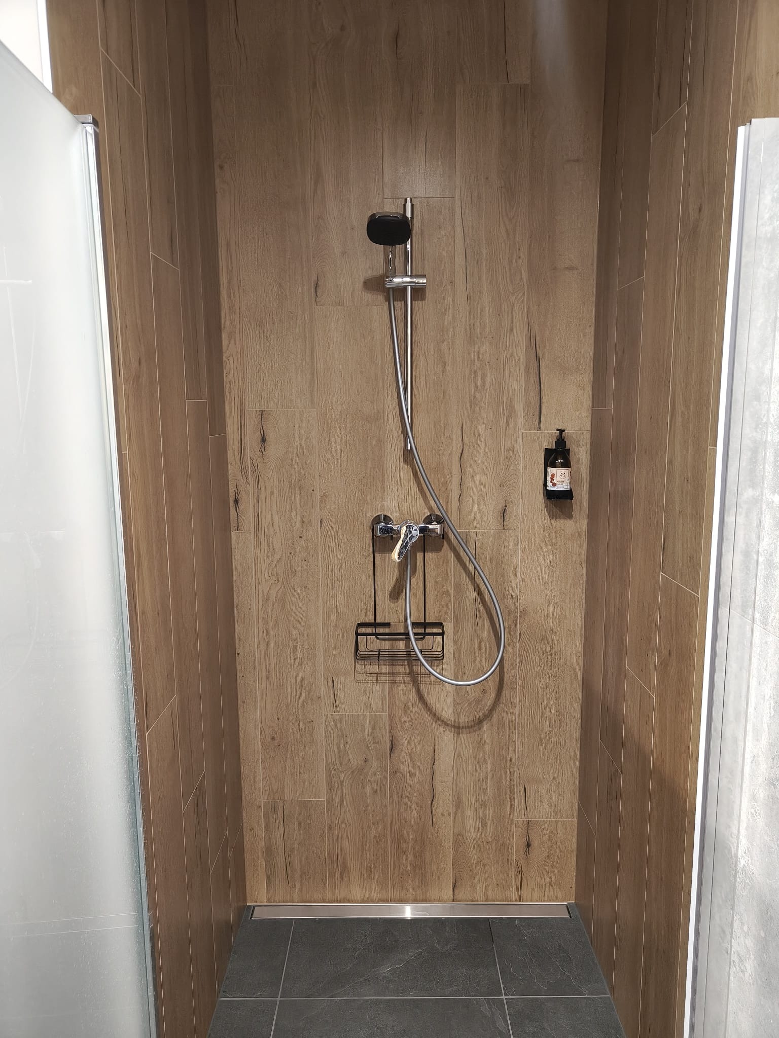 Kabina prysznicowa z deszczownicą i półką na kosmetyki, wykończona płytkami imitującymi drewno. Szara podłoga i szklane drzwi kabiny.