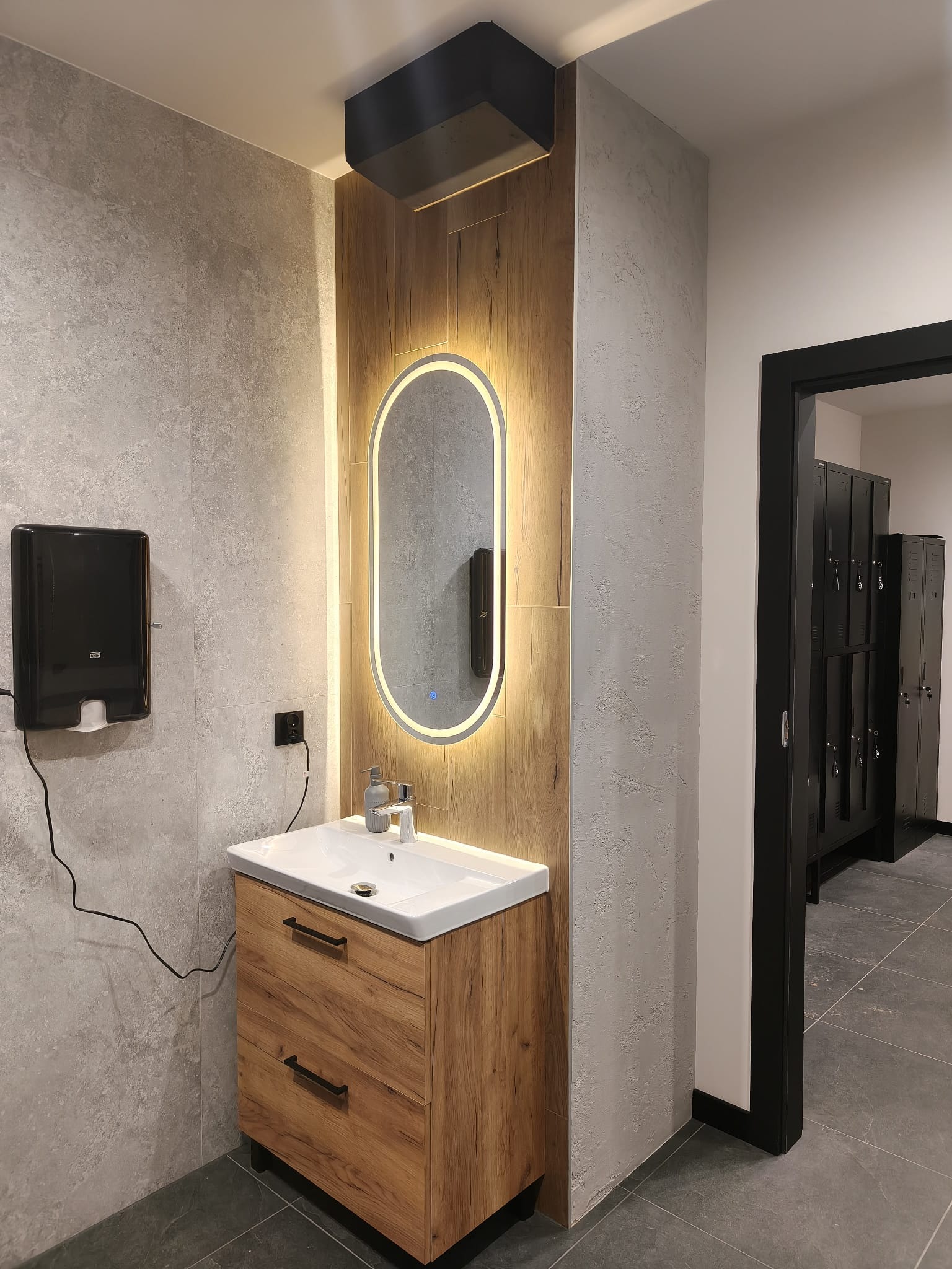 Nowoczesna łazienka z drewnianą szafką pod umywalką, podświetlanym lustrem i szarą płytką. Widoczne czarne szafki w tle. Minimalistyczny design.