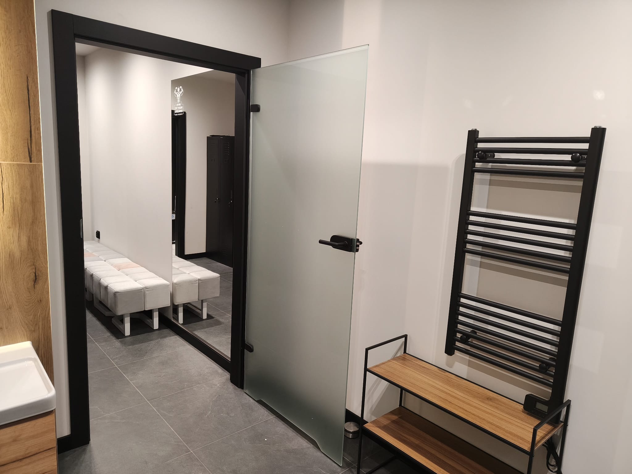 Nowoczesna łazienka z szarą podłogą, szklanymi drzwiami i czarnym grzejnikiem drabinkowym. Minimalistyczny design z elementami drewna.