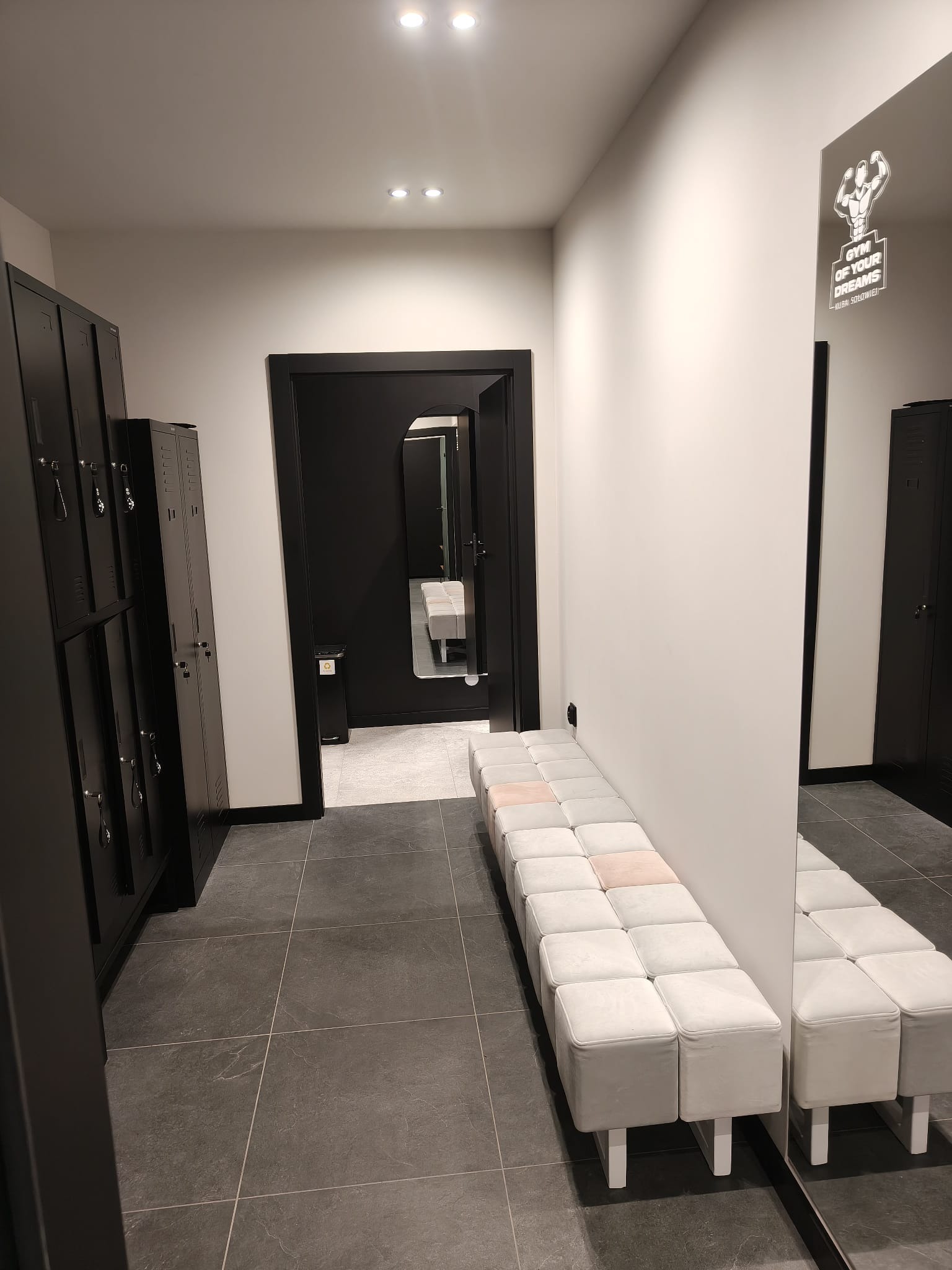Nowoczesna szatnia w siłowni: czarne szafki, szara podłoga, biała ławka z pufami i lustro z logo 'Gym of Your Dreams'. Minimalistyczny design, oświetlenie punktowe.