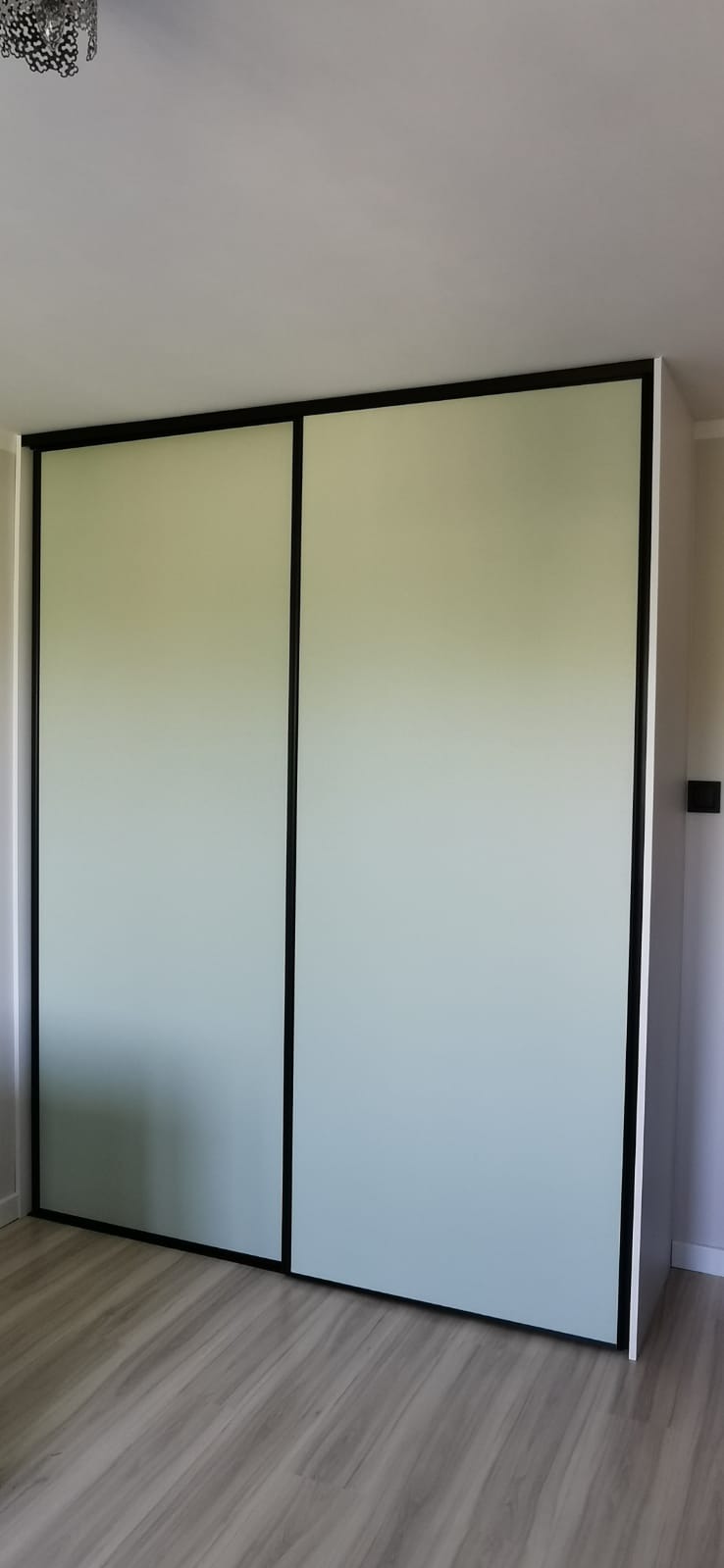 Nowoczesna szafa przesuwna z jasnymi frontami i czarnymi obramowaniami, dopasowana do wnętrza z panelami podłogowymi. Minimalistyczny design.