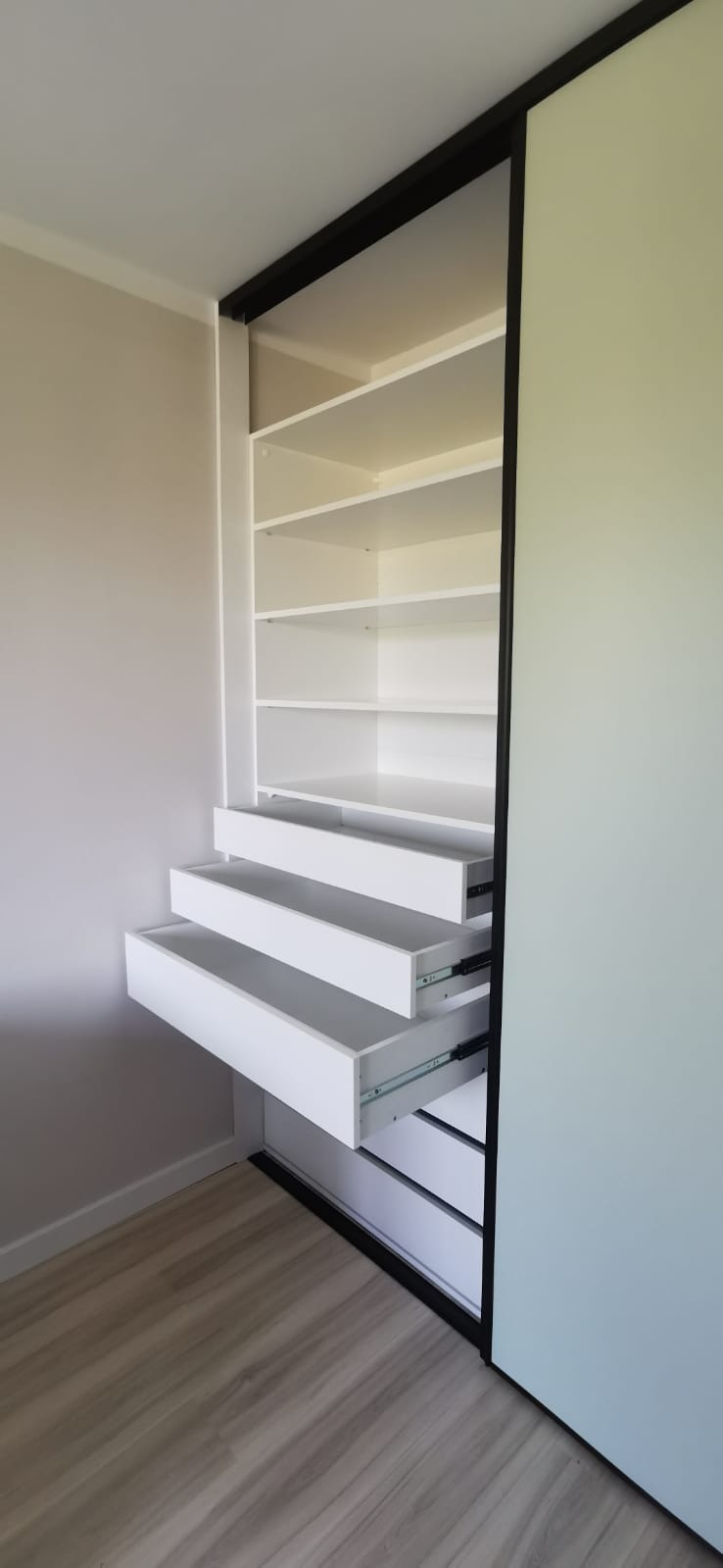 Biała szafa wnękowa z otwartymi szufladami i półkami, czarne obramowanie drzwi przesuwnych, widok z dołu, jasne wnętrze.