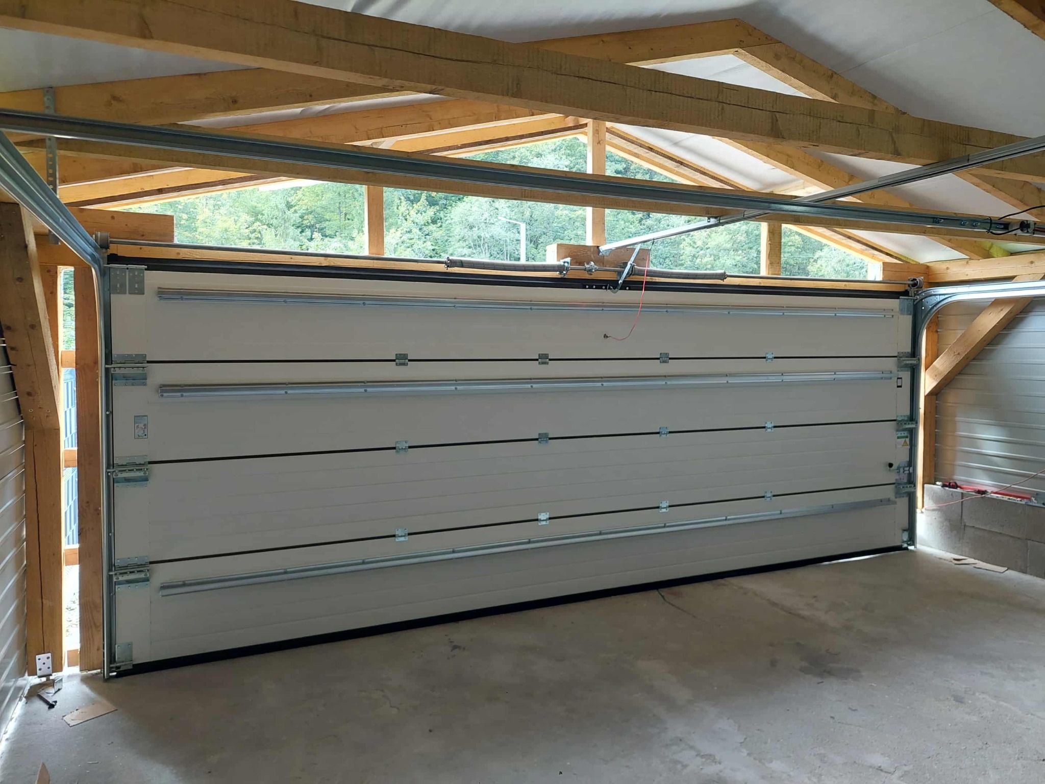 Zamknięta segmentowa brama garażowa w jasnym odcieniu, widoczna od wewnątrz garażu z drewnianą konstrukcją dachu i fragmentem otoczenia za nią.