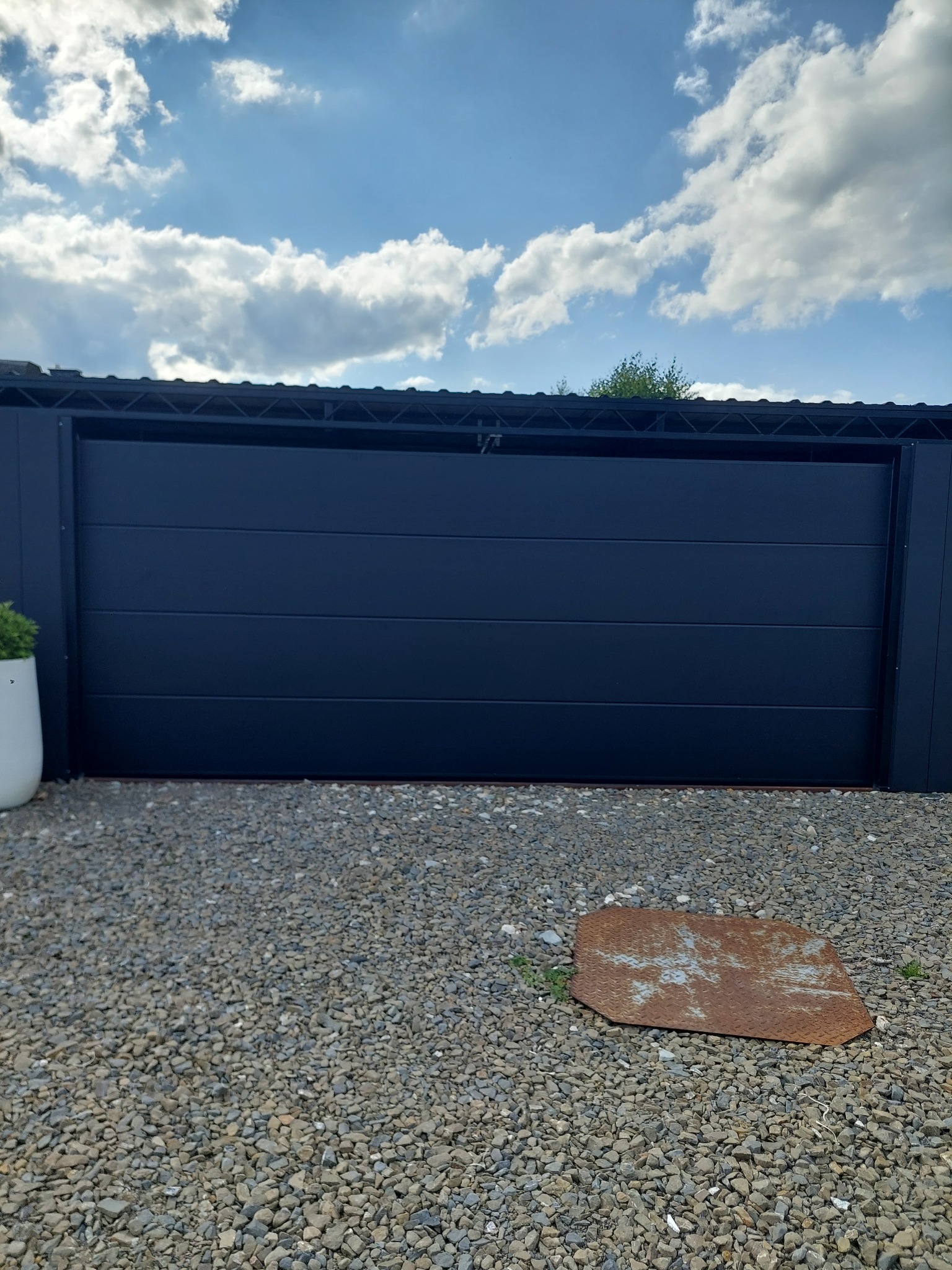 Nowoczesna, przesuwna brama w kolorze antracytu, z widoczną fakturą poziomych paneli. Na pierwszym planie żwirowe podłoże i metalowa płyta. W tle błękitne niebo z chmurami.