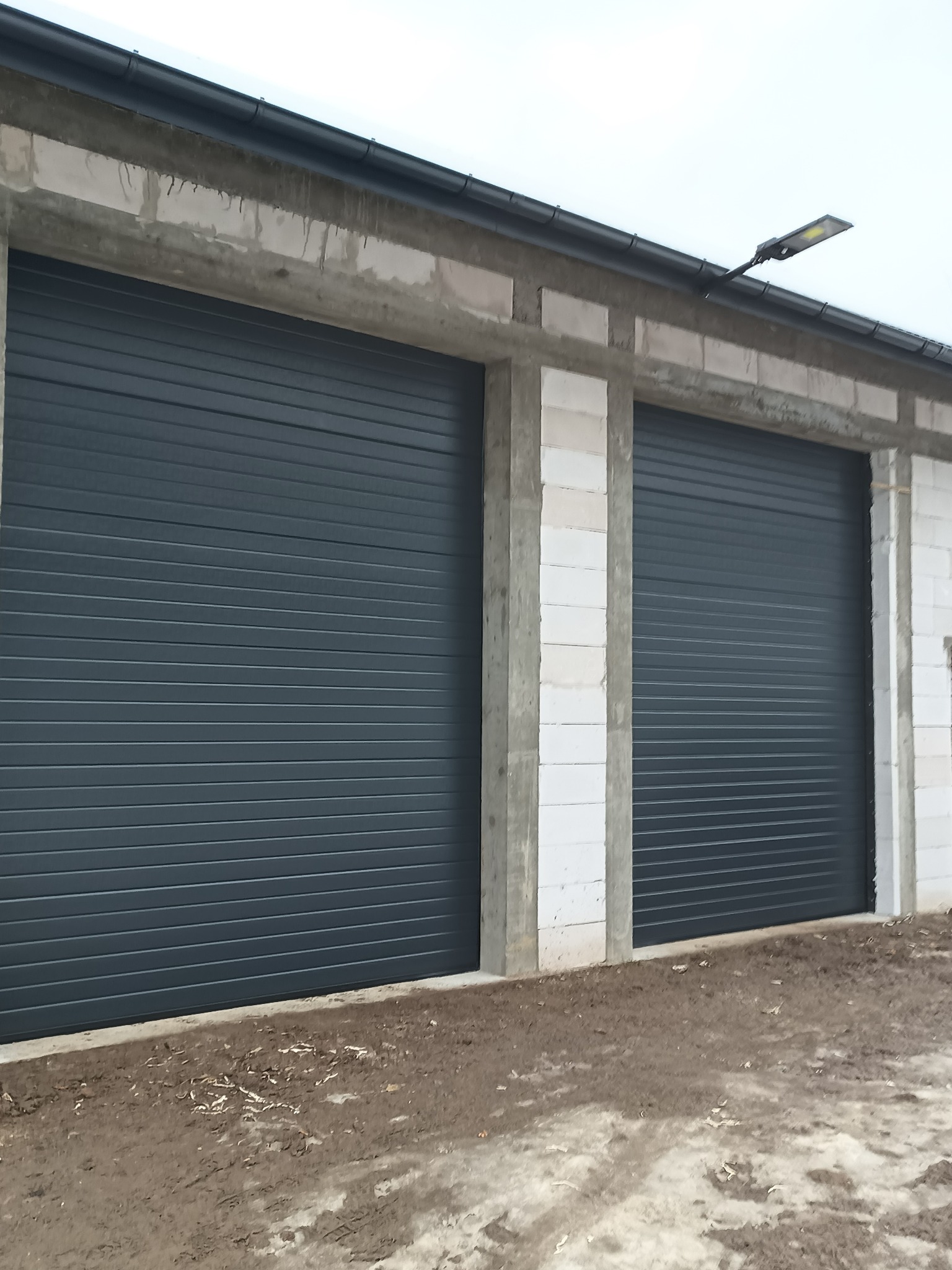 Dwie szare bramy garażowe segmentowe w surowym budynku z betonu komórkowego, oświetlone lampą zewnętrzną. Widok z zewnątrz, na pierwszym planie ziemia.