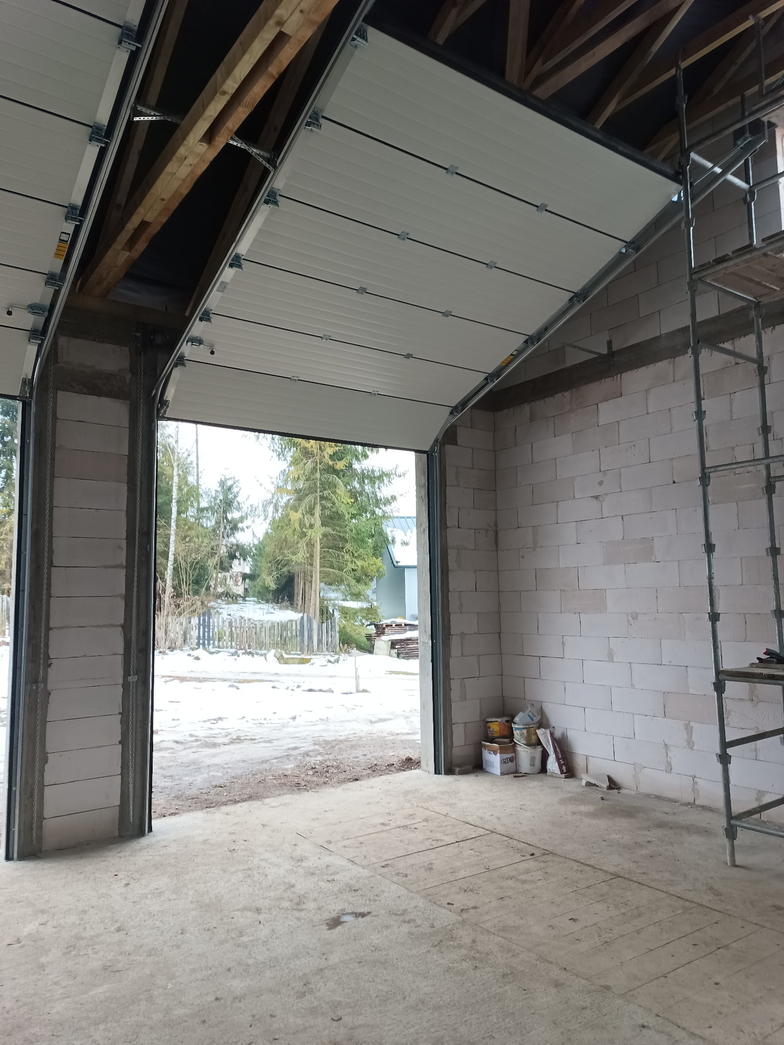 Montaż segmentowej bramy garażowej w surowym garażu z bloczków, widoczne elementy konstrukcyjne i rusztowanie. Zima za oknem.