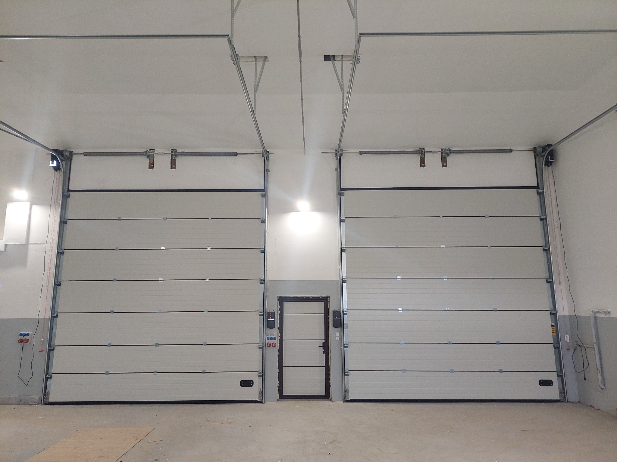 Dwie segmentowe bramy garażowe w jasnym pomieszczeniu, z drzwiami przejściowymi pośrodku. Widoczne prowadnice i sprężyny bram, oraz instalacja elektryczna po bokach.