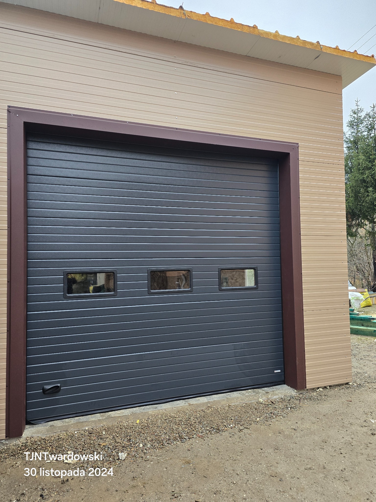 Szara brama garażowa segmentowa z trzema małymi okienkami, brązowe obramowanie, beżowa elewacja budynku. Widok z zewnątrz, brama zamknięta.