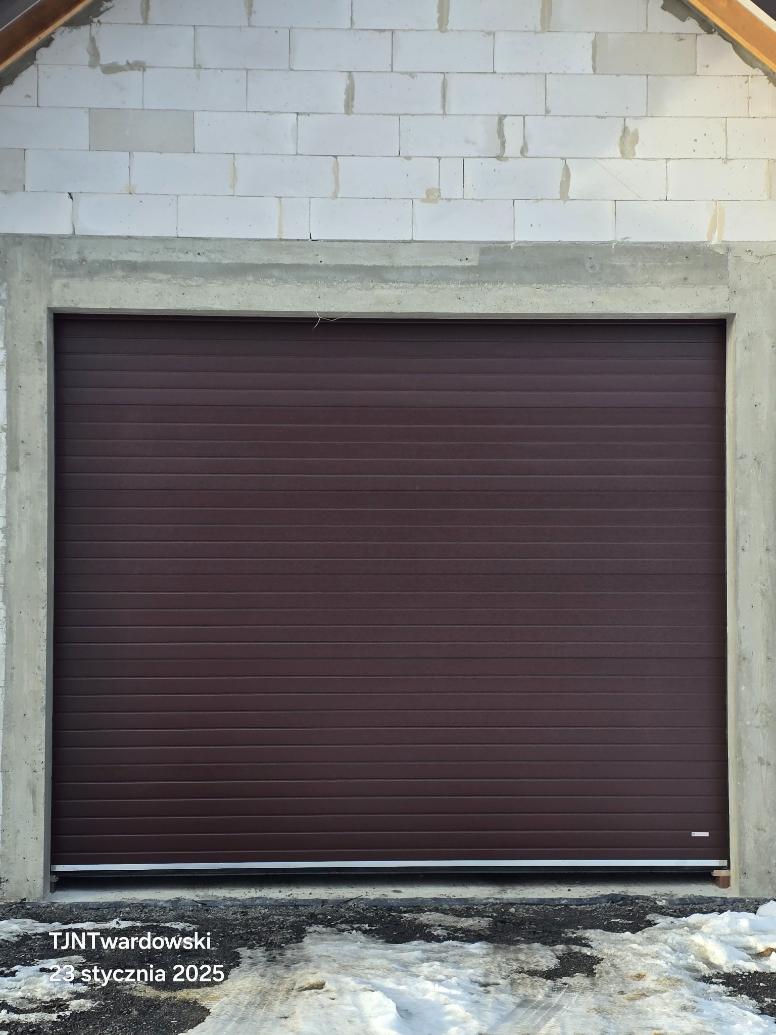 Zamknięta, brązowa brama garażowa segmentowa w surowym otoczeniu betonowej ramy i muru z bloczków. Na dole widoczny śnieg i błoto.