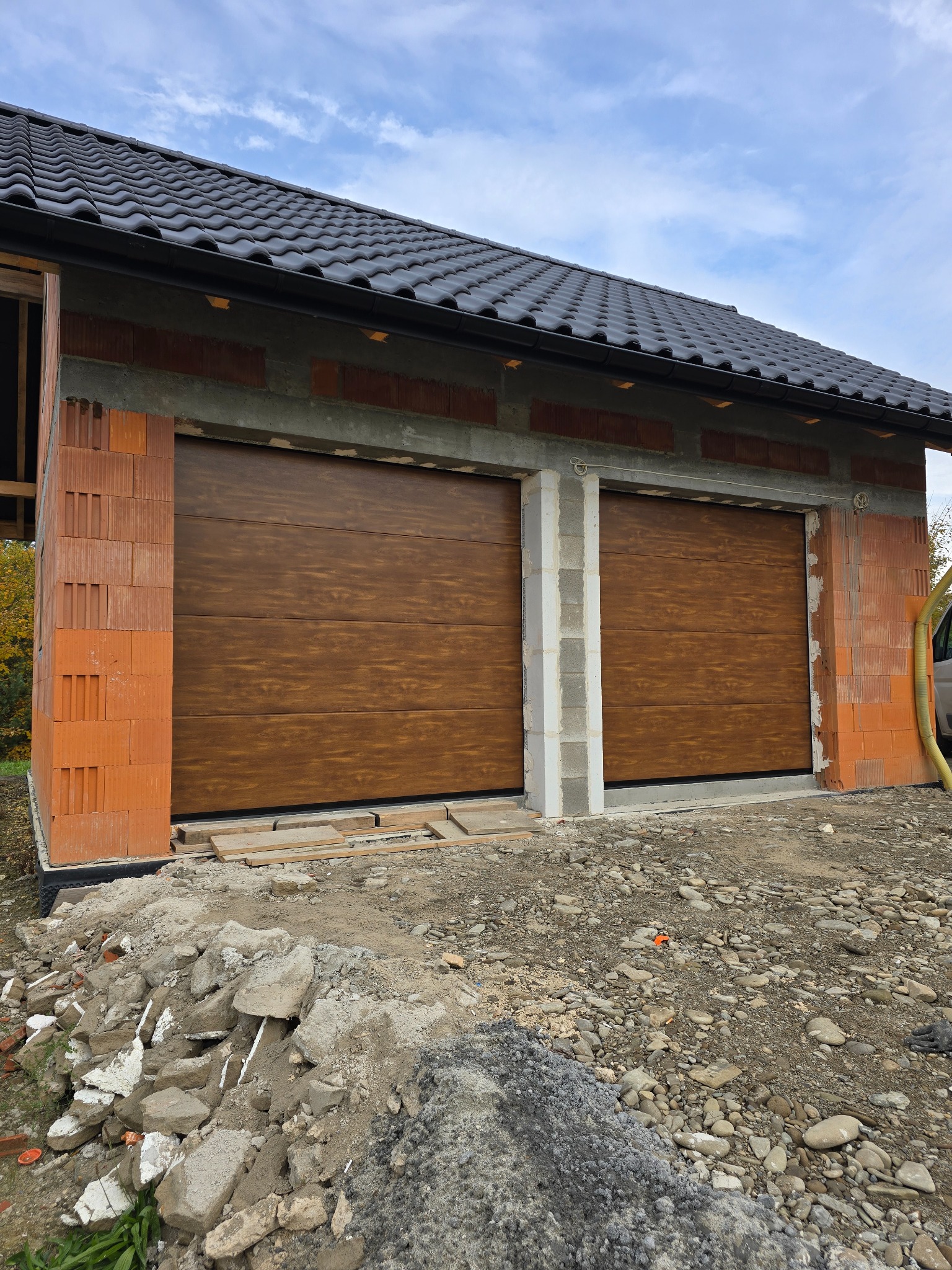 Dwie brązowe bramy garażowe segmentowe, zamontowane w surowym budynku z cegły, z widocznym gruzem budowlanym na zewnątrz. Dach pokryty czarną dachówką.